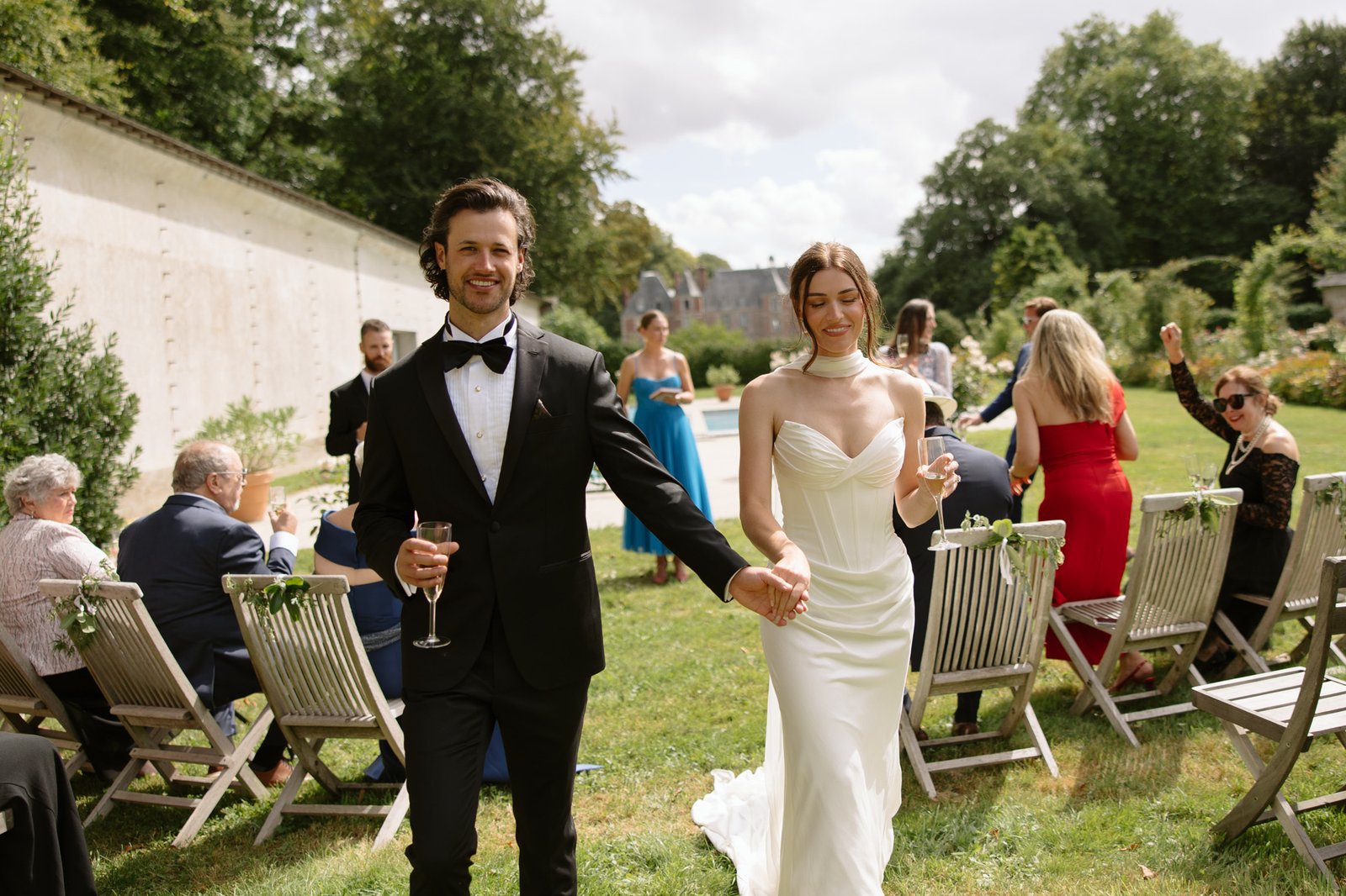 A bride and groom walk hand in hand outdoors, smiling, with wedding guests seated on wooden chairs in the background. Normandy Chateau Wedding.