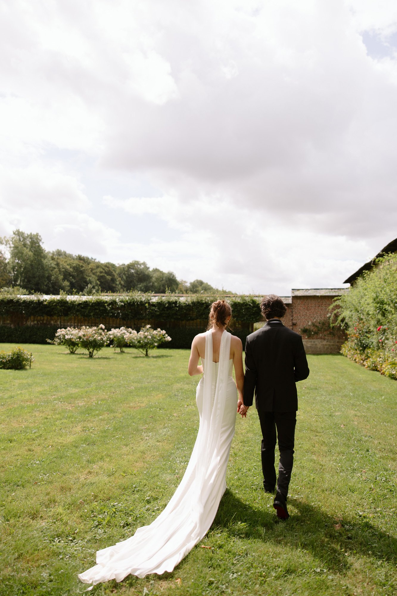 A bride in a white dress and a groom in a black suit walk hand in hand across a grassy garden, seen from behind.