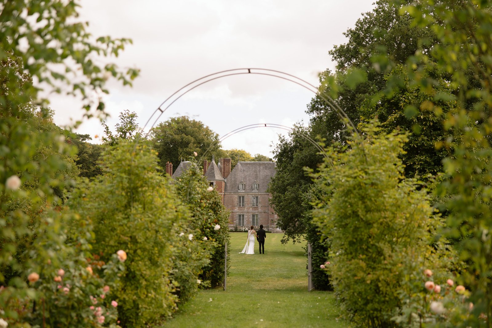 A bride and groom walk together along a green garden path toward a large stone house, surrounded by trees and archways. Normandy Chateau Wedding.