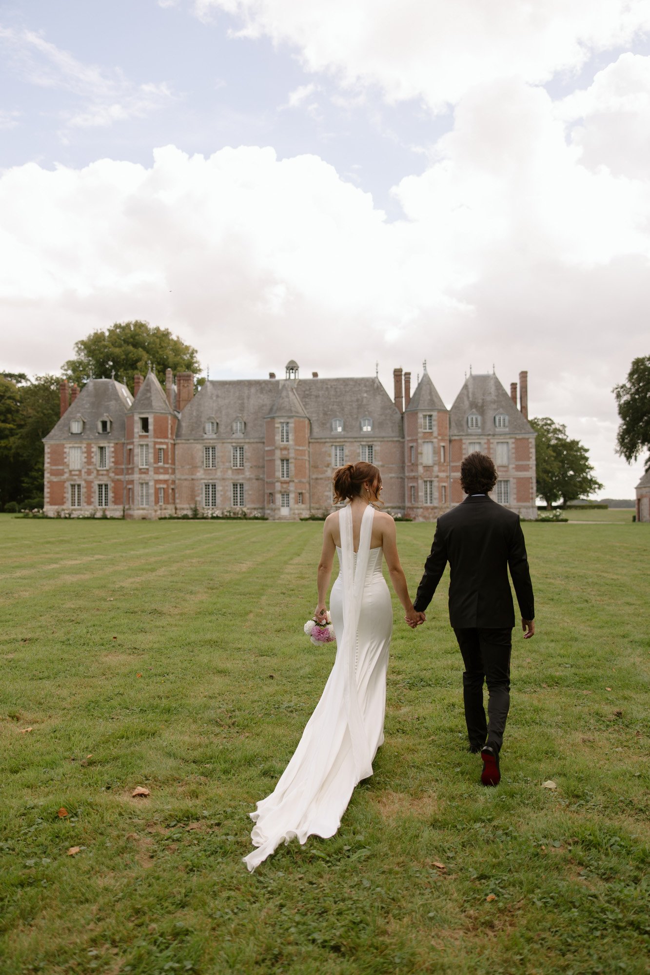 A couple in formal attire walks hand in hand across a lawn toward a large historic mansion under a cloudy sky.