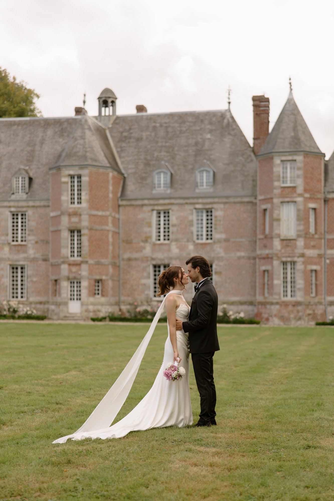 A bride and groom stand together on a lawn in front of a large historic building, facing each other. The bride holds a bouquet and wears a long white dress with a cape. Normandy Chateau Wedding.