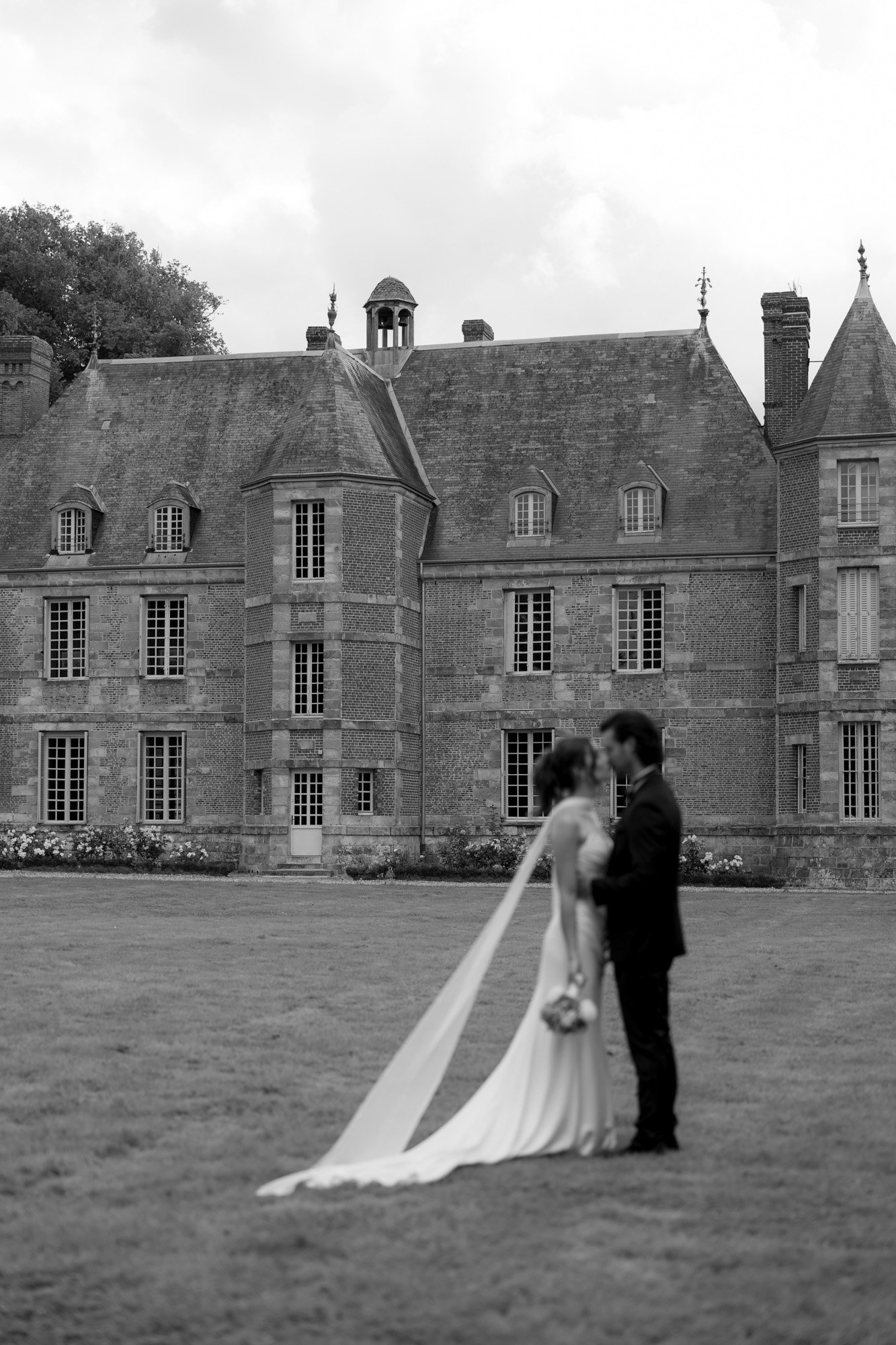 A bride and groom stand closely together on a lawn in front of a large, historic building; the background is in focus while the couple is blurred.