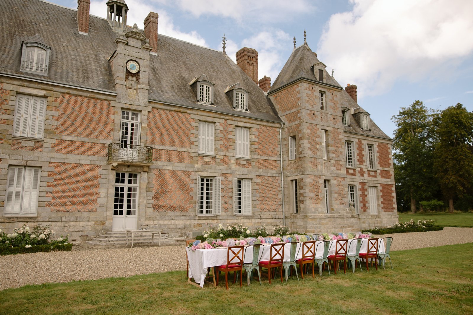 A long outdoor dining table with chairs is set up on the lawn in front of a large, historical brick and stone building.