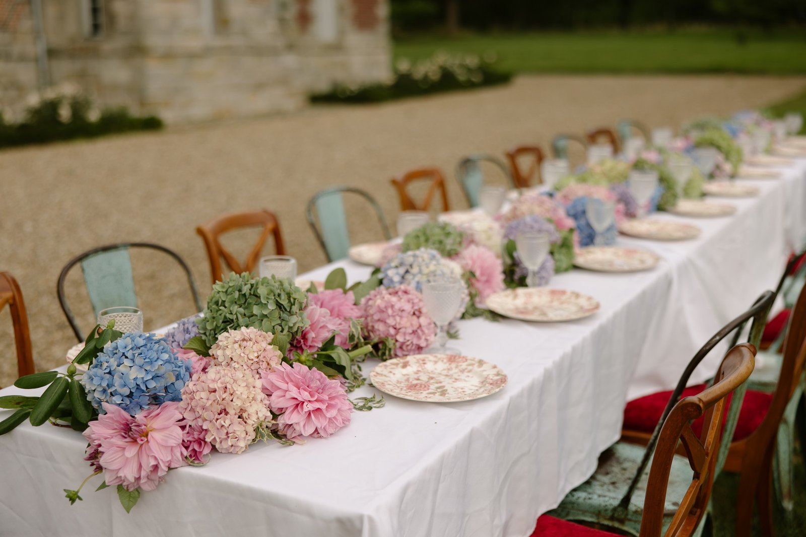 A long outdoor table with a white cloth, floral plates, and pastel hydrangea centerpieces, surrounded by mixed wooden chairs.