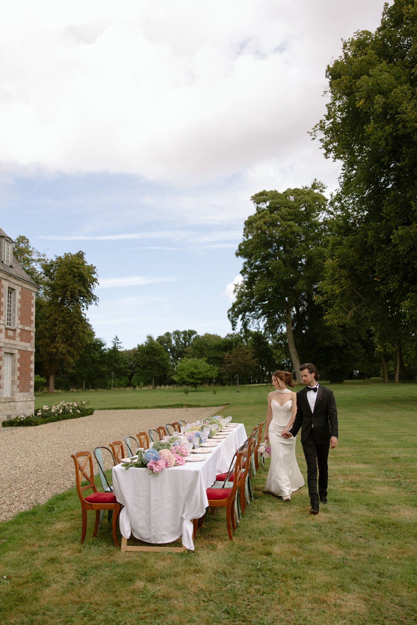 A bride and groom walk past a long outdoor table set for a formal meal on the lawn of a large estate with trees and gardens in the background. Normandy Chateau Wedding.