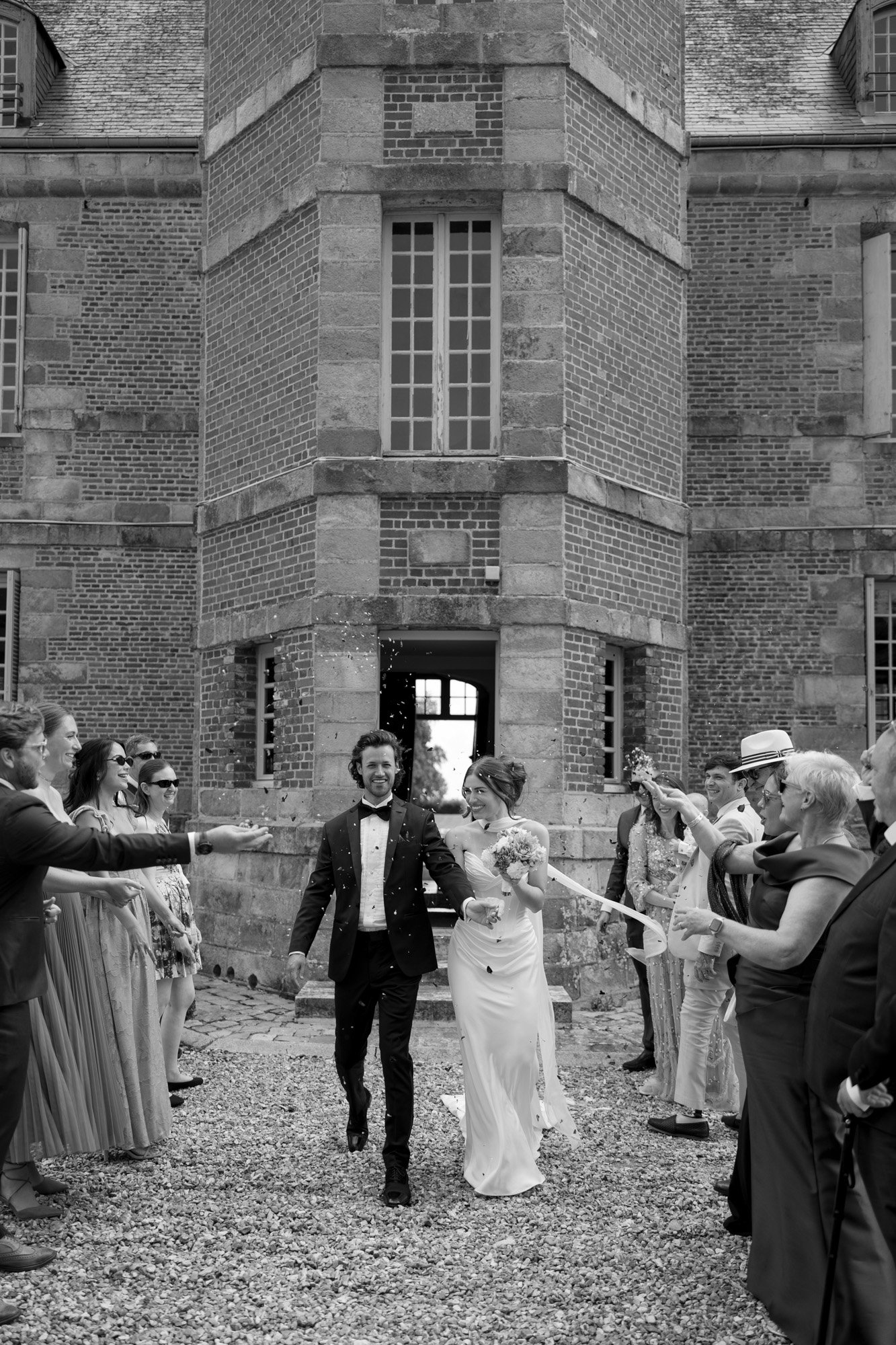 A bride and groom walk outside a brick building as guests on both sides throw confetti and celebrate their wedding.