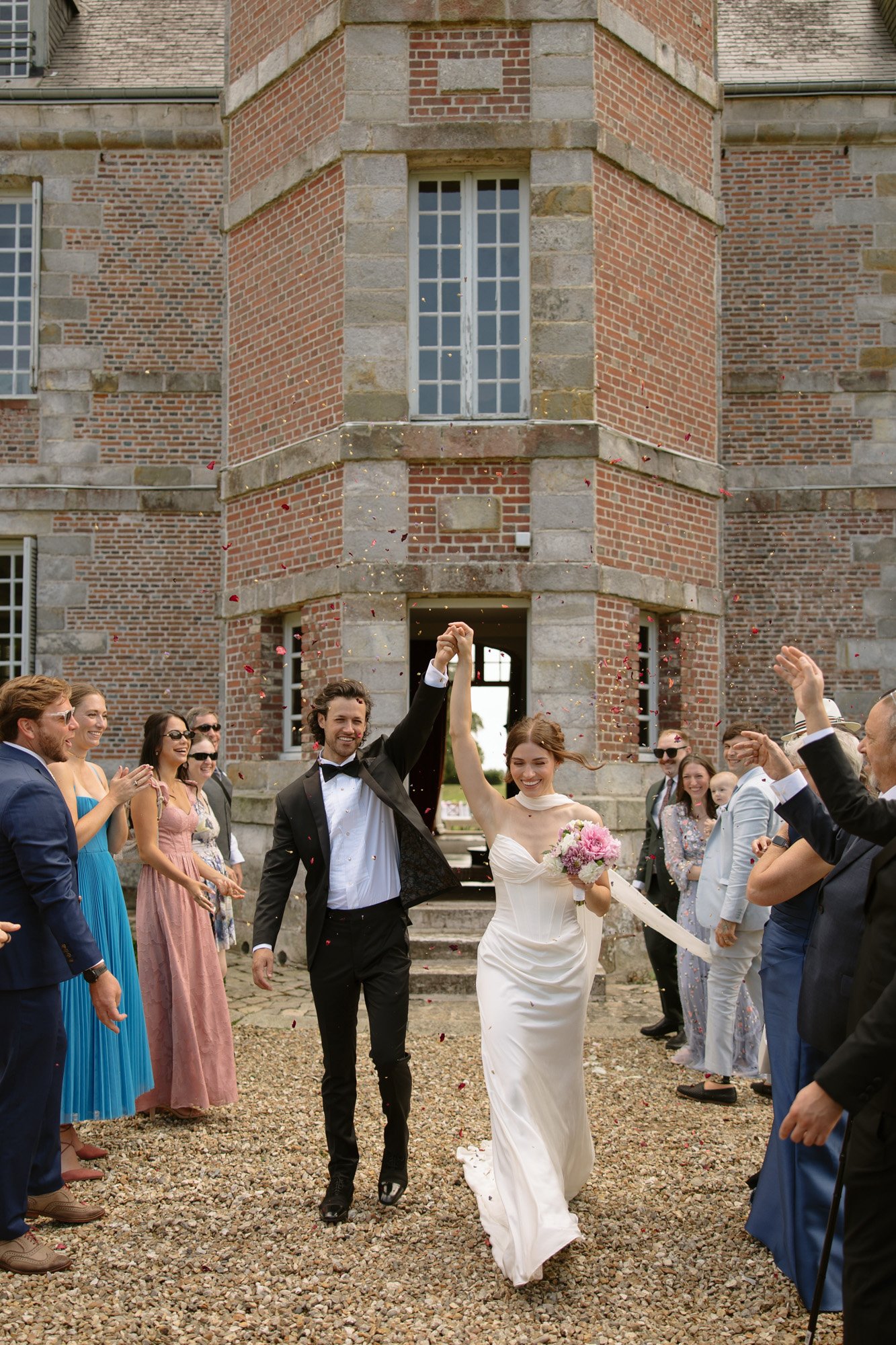A bride and groom walk hand in hand outside a brick building as wedding guests stand on either side, throwing flower petals in celebration. Normandy Chateau Wedding.