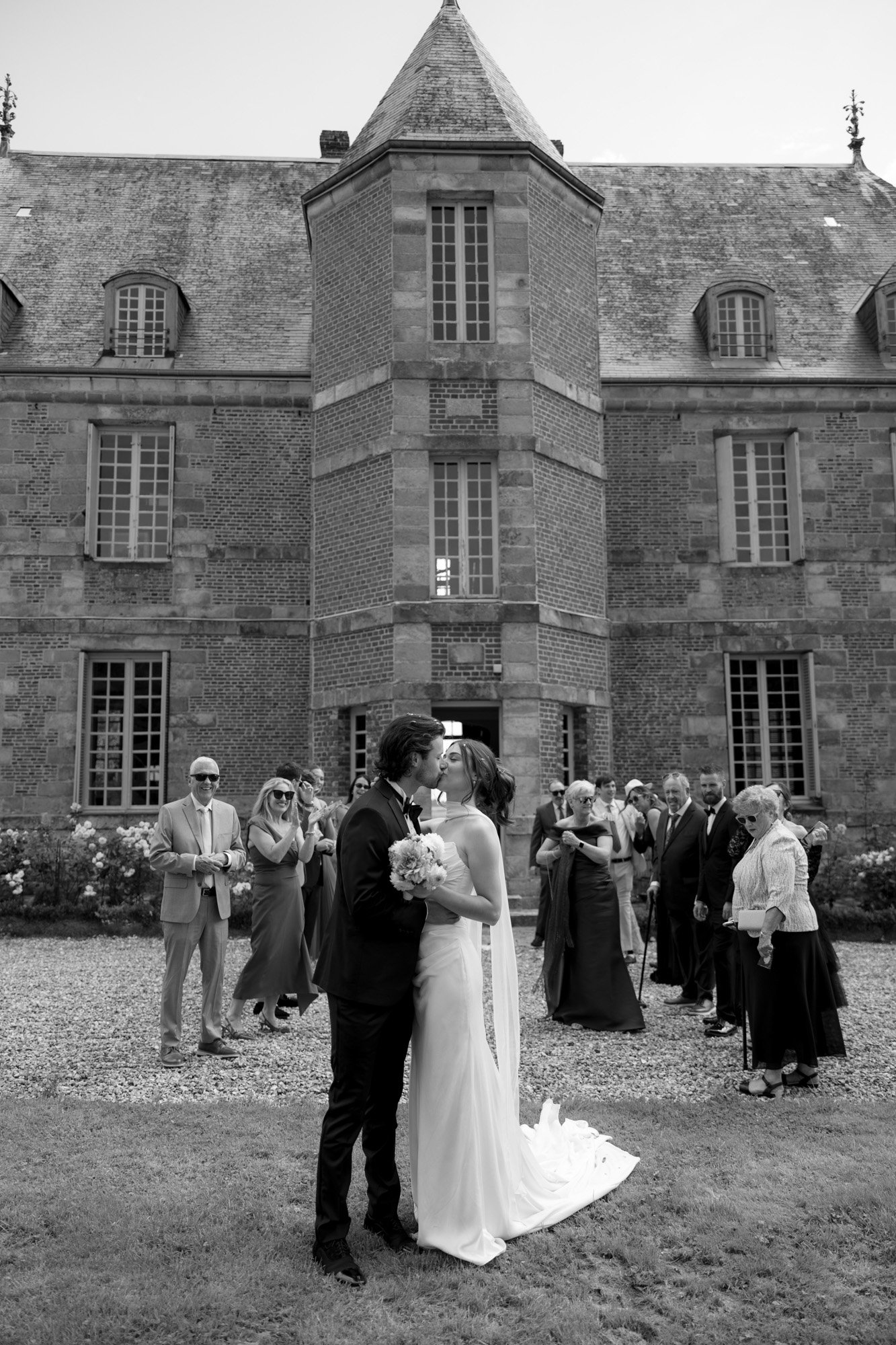 A bride and groom kiss in front of a historic building as wedding guests stand and watch, some taking photos. Normandy Chateau Wedding.