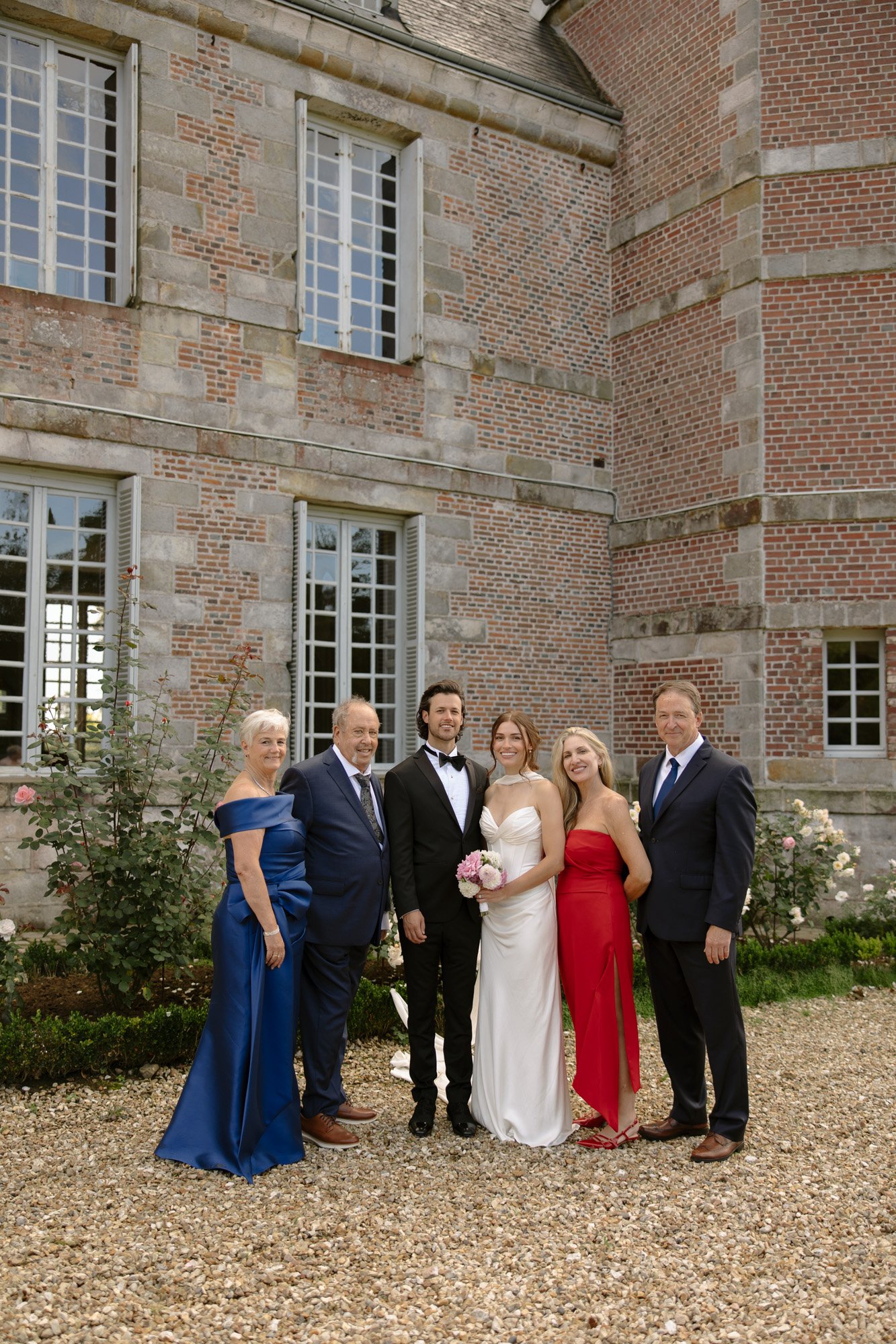 Six people dressed formally, including a bride and groom, stand together outside a brick and stone building, posing for a group photo.
