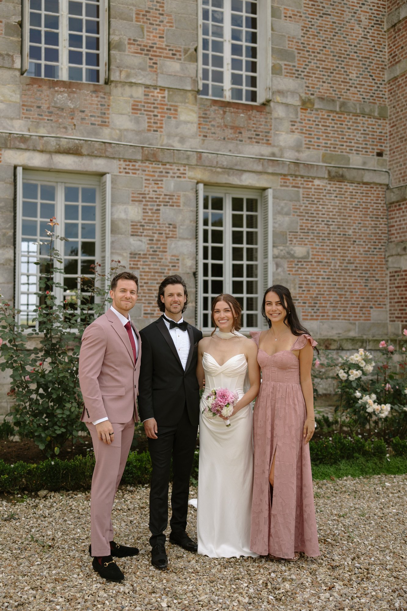 Four people pose outdoors in formal attire in front of a brick building with large windows; two men in suits and two women in dresses, one holding a bouquet.