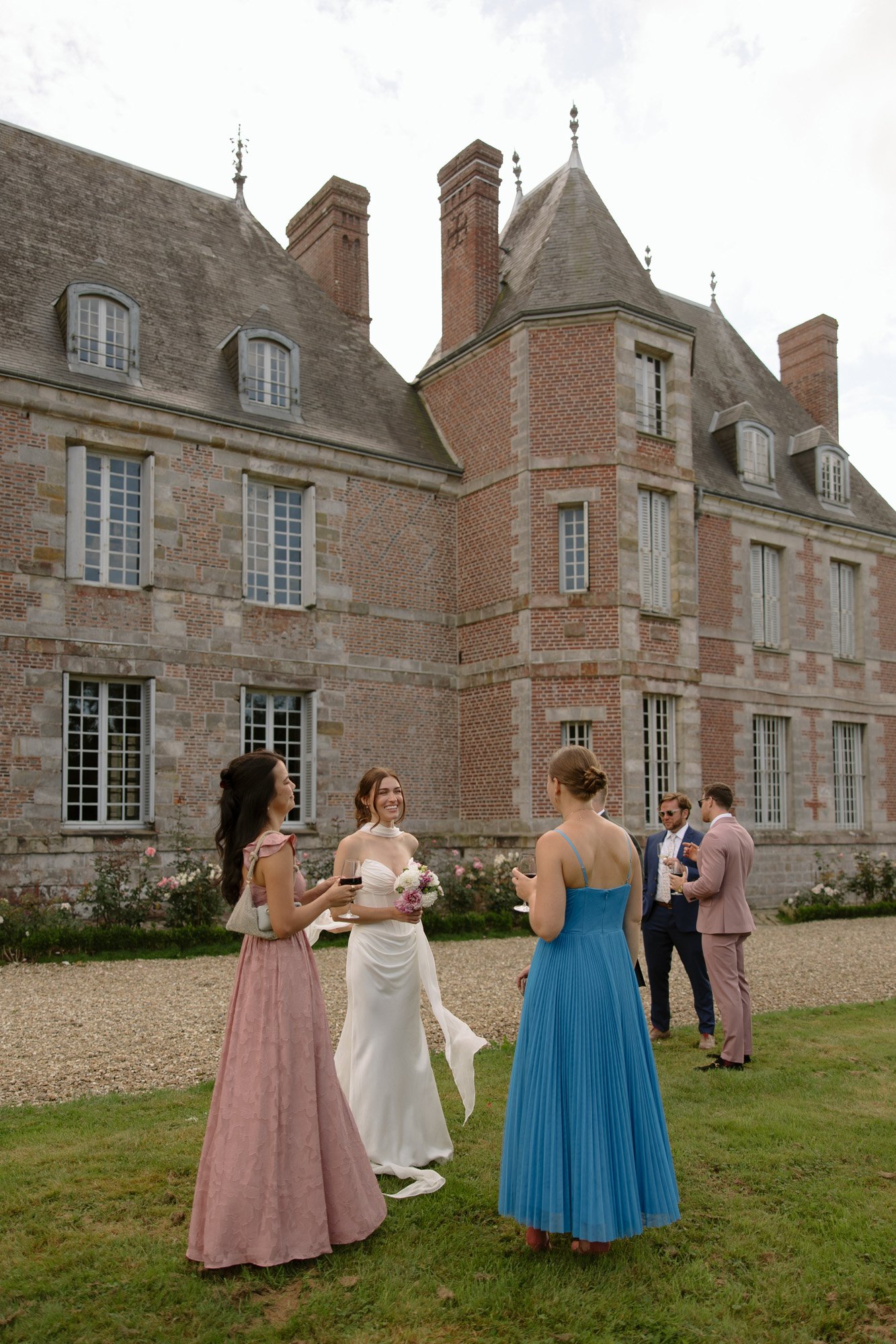 A bride in a white dress stands on a lawn with three guests in formal dresses, with a historic stone and brick building in the background. Normandy Chateau Wedding.