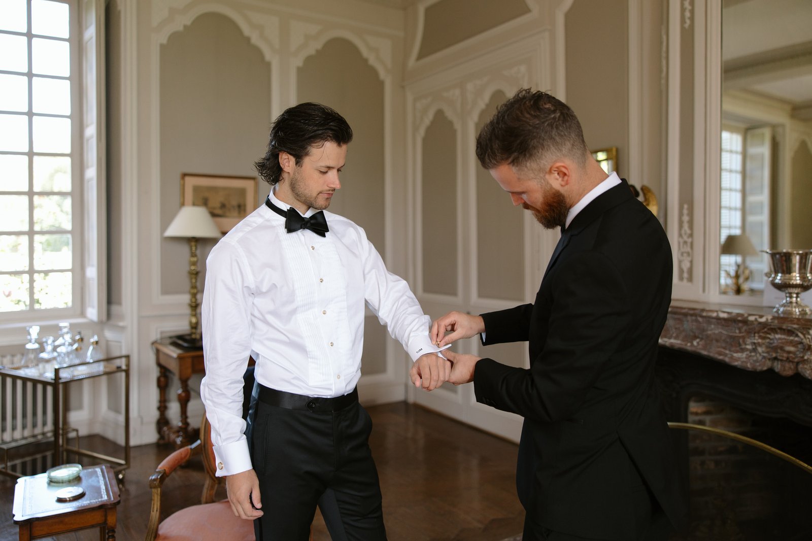 Two men in formal attire stand in an elegant room; one adjusts the other’s shirt cuff as they prepare, possibly for a formal event or ceremony.