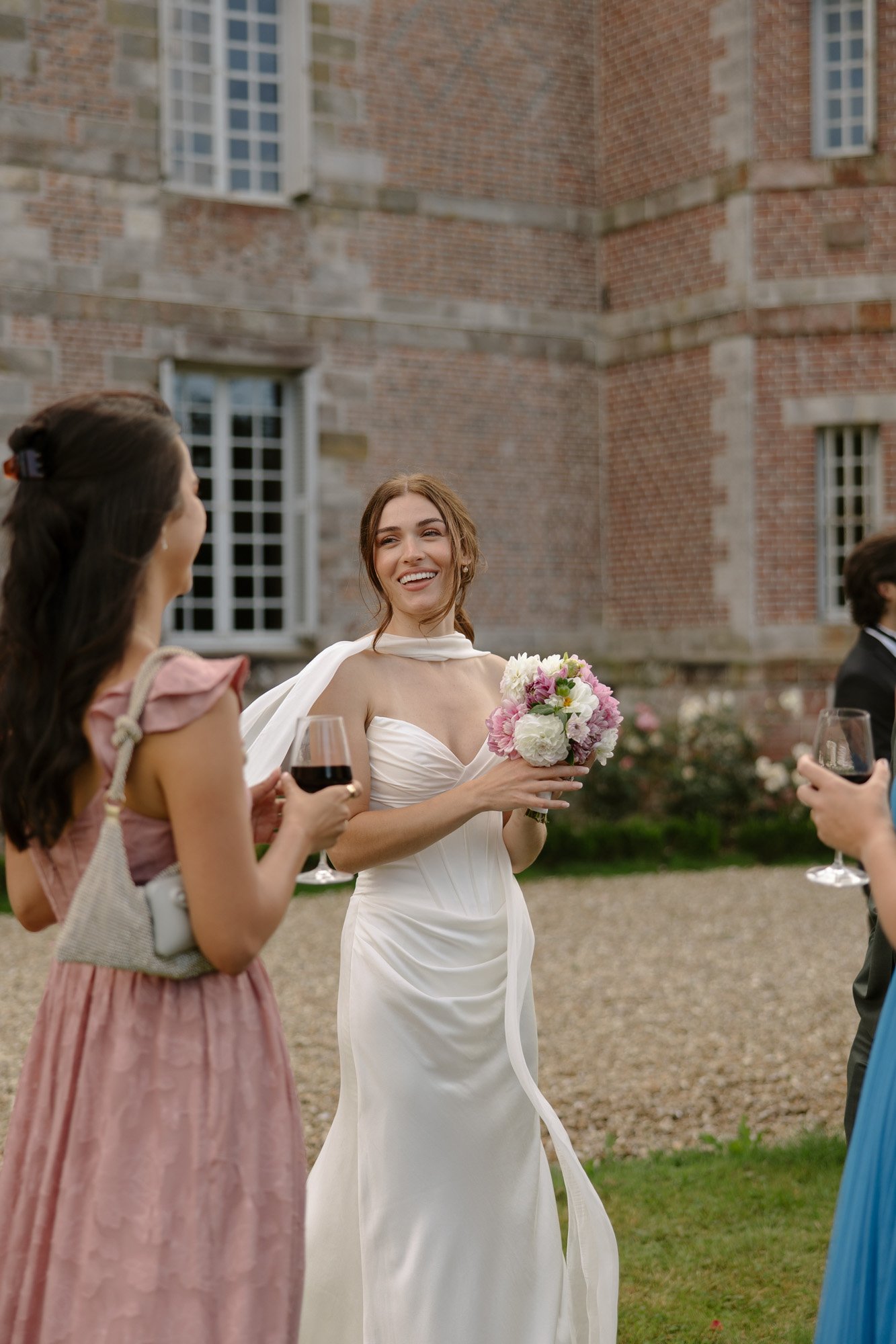 A bride in a white dress holds a bouquet and smiles while talking to guests at an outdoor event near a brick building.
