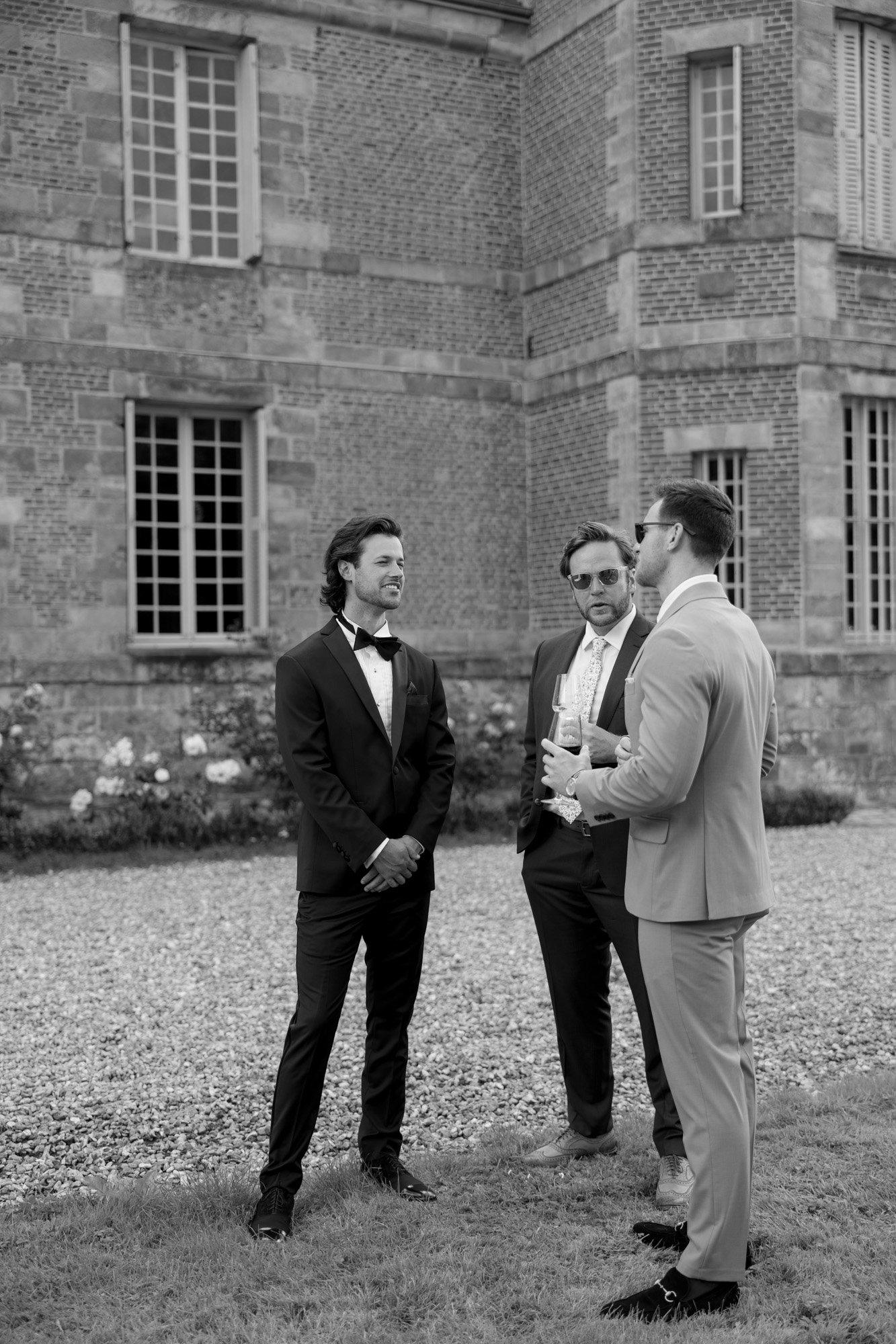 Three men in suits stand and talk outside a brick building on a gravel path, with one man holding a drink.