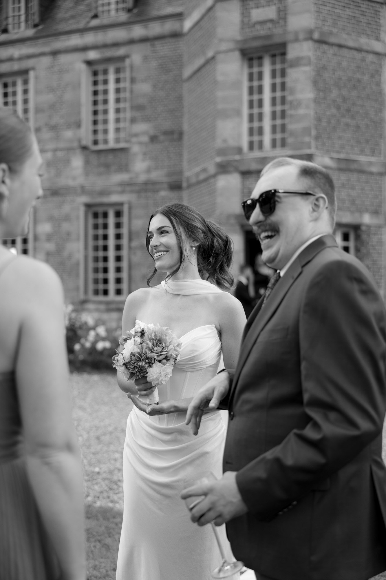 A bride holding a bouquet smiles and talks with two guests, one man in sunglasses and a suit, outside a large brick building.
