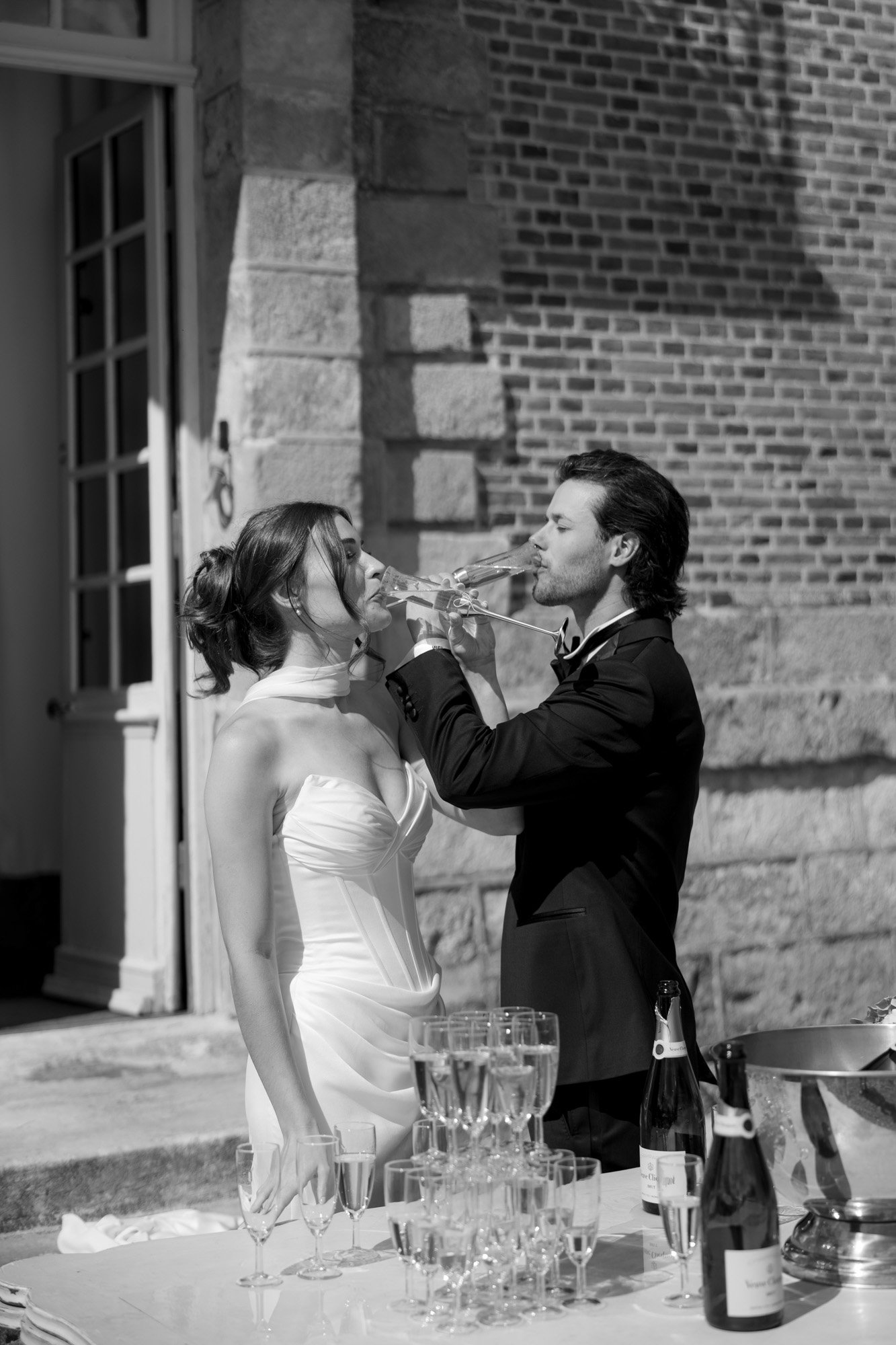 A couple dressed in formal attire drink from champagne glasses outside near a table with bottles and empty glasses.