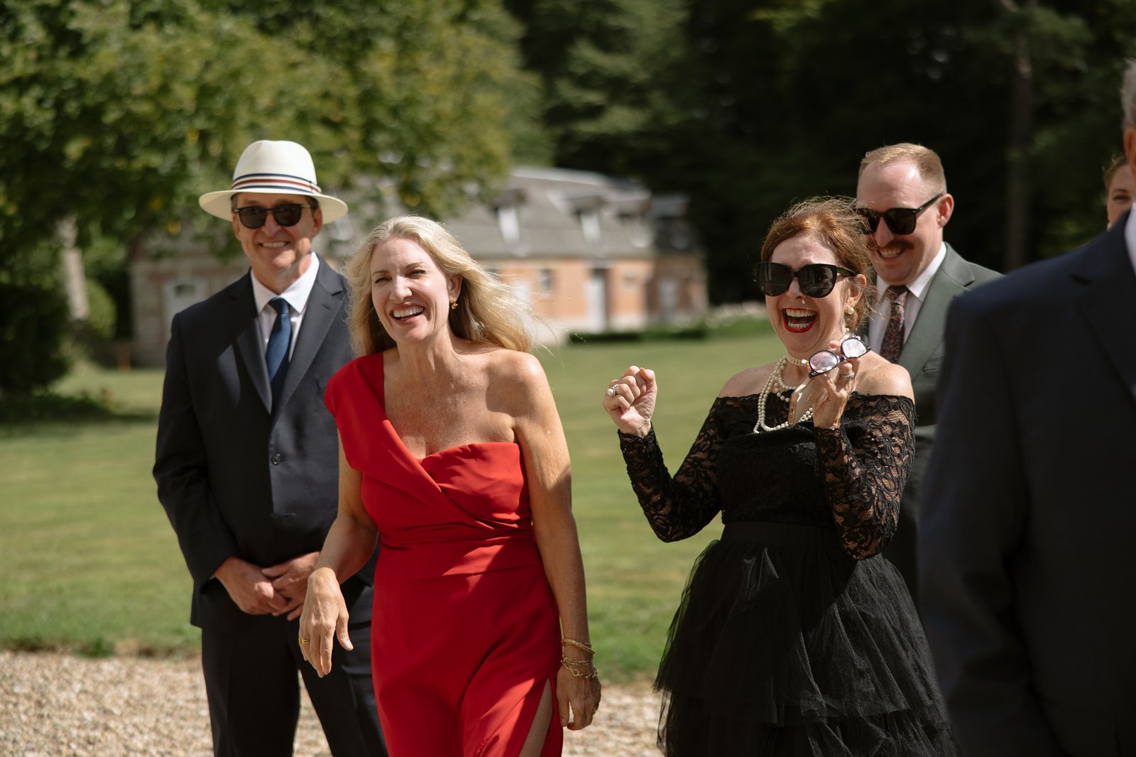 Four adults dressed formally smile and laugh outdoors on a sunny day, with green grass and buildings in the background.