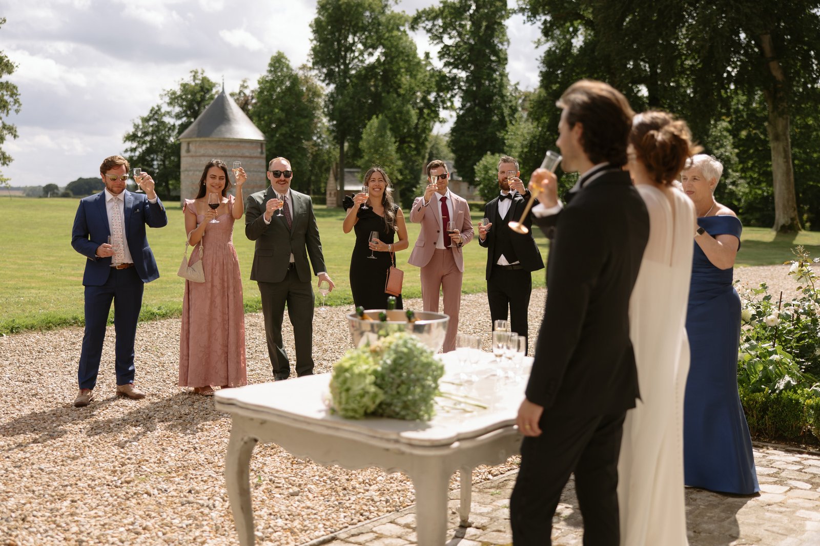 A group of people dressed formally stand outdoors, raising glasses in a toast toward a couple at a table with drinks and a flower arrangement.