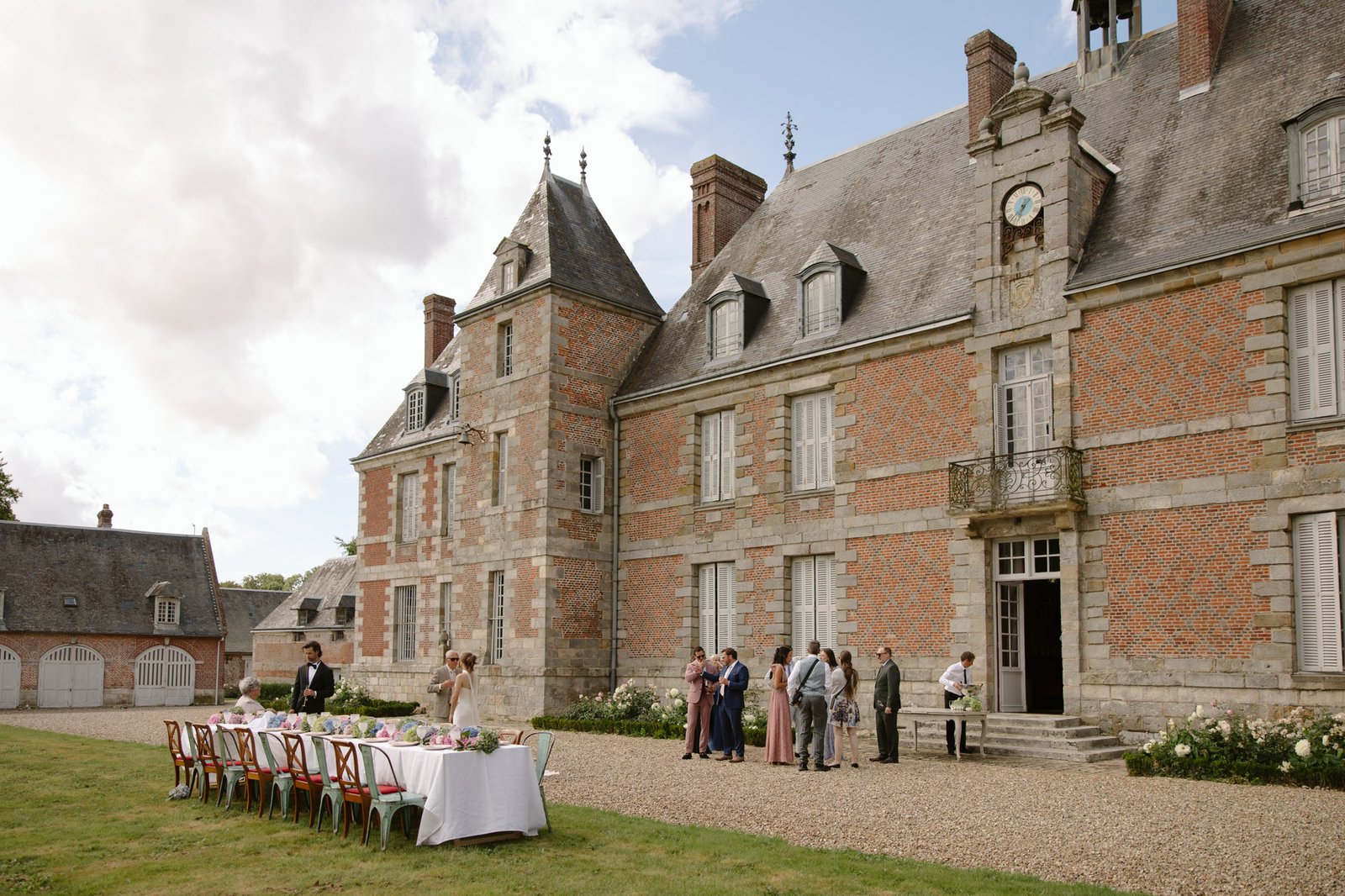 A group of people gather outside a large historic brick building with a clock, near a long outdoor dining table set for a meal. Normandy Chateau Wedding.