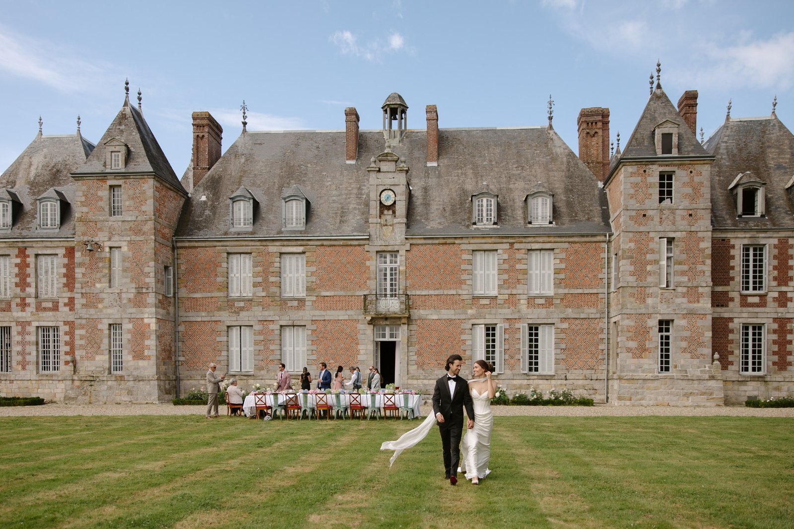A couple in formal attire walks across a lawn in front of a large historic chateau, with a group of people seated at a long outdoor table in the background. Normandy Chateau Wedding.