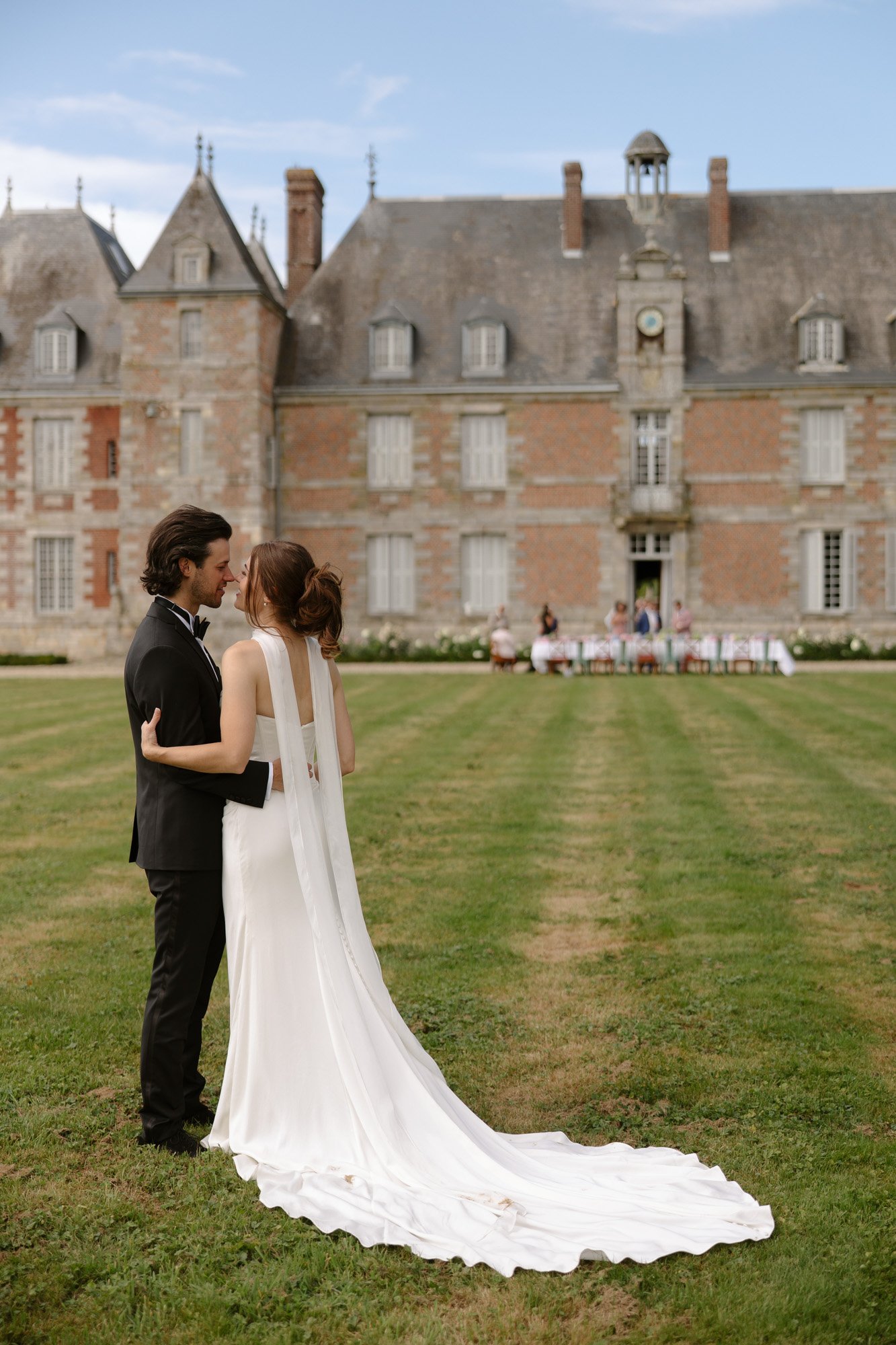 A bride and groom stand on a lawn facing each other, with a large historic building and seated guests visible in the background.