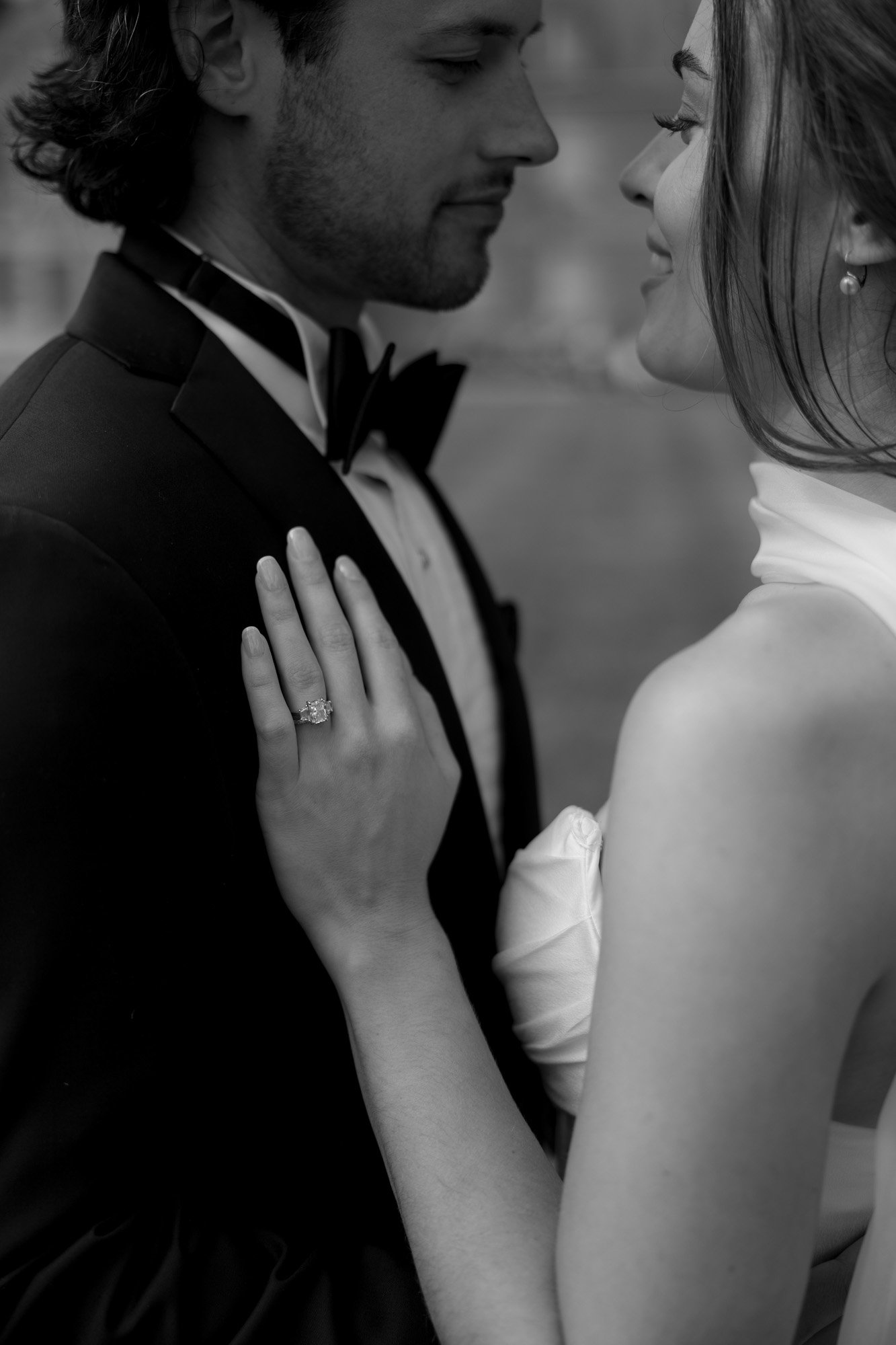 A woman in a white dress with an engagement ring rests her hand on a man’s chest; both are dressed formally and facing each other closely.