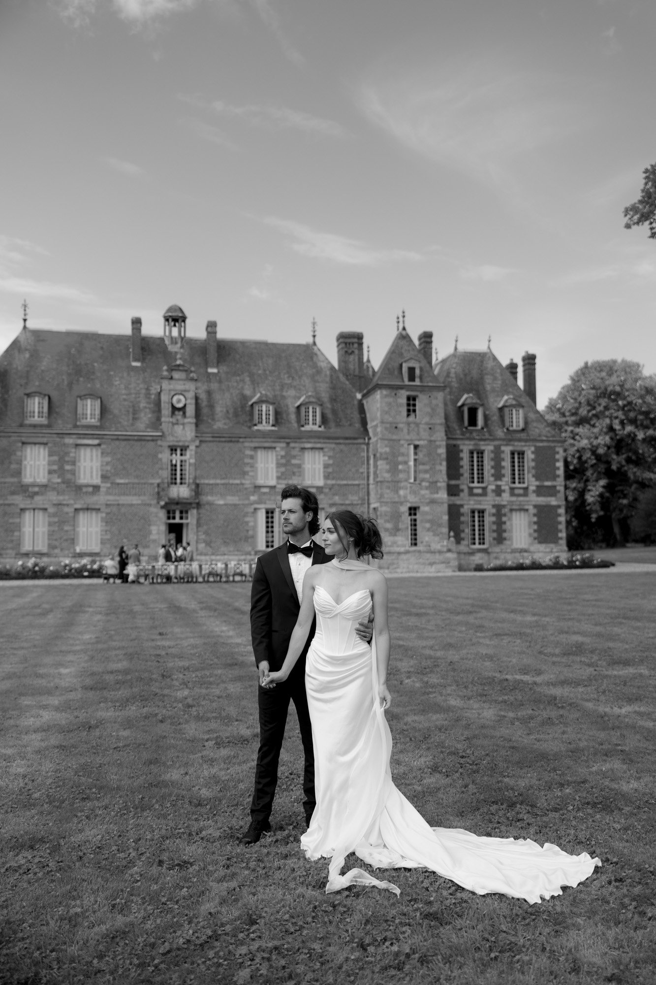 A bride and groom stand together on a lawn in front of a large historic building, both dressed formally; the scene is in black and white. Normandy Chateau Wedding.