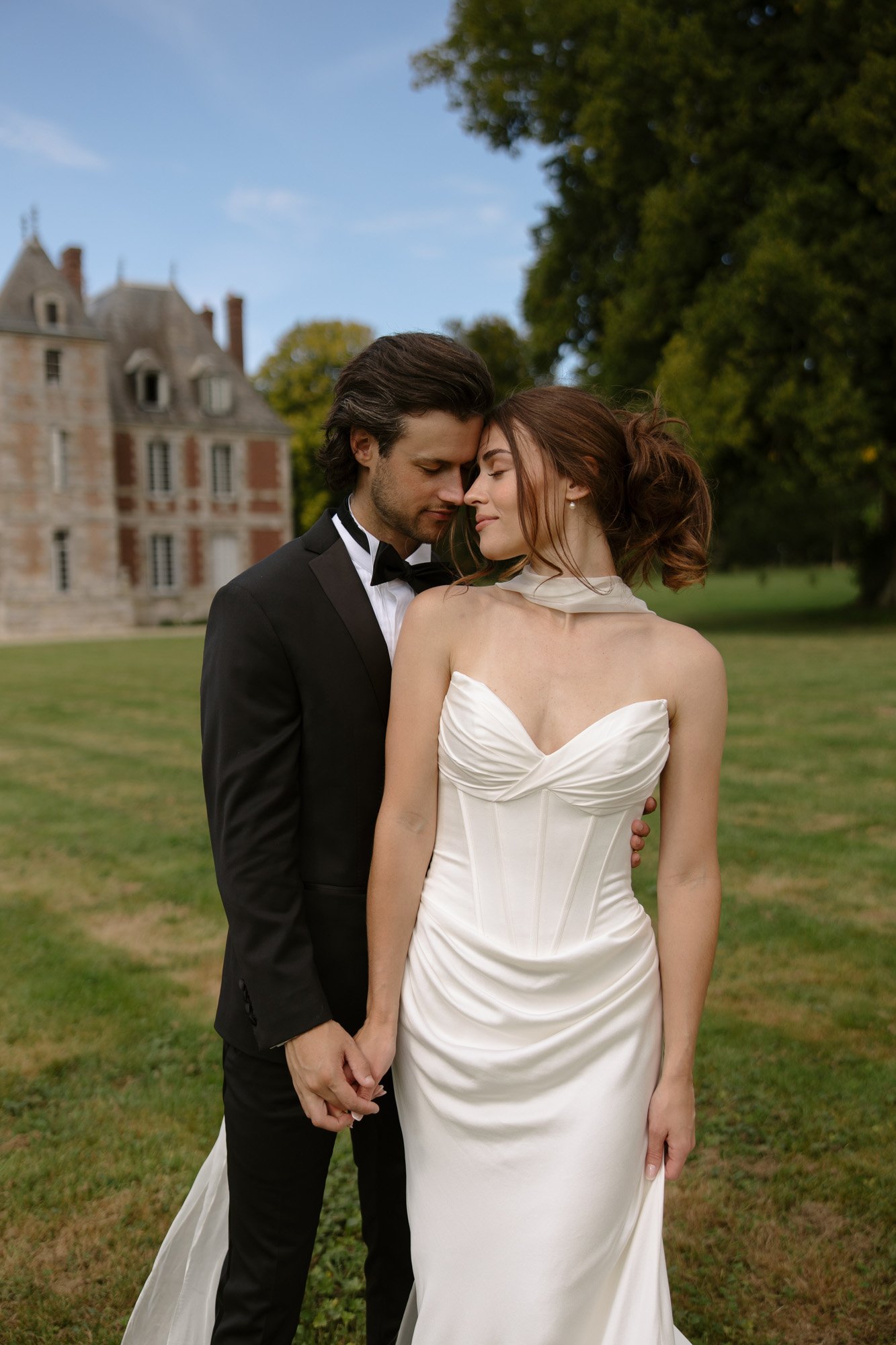 A couple dressed in formal attire stands close together on a lawn, with a historic stone building and trees in the background. Normandy Chateau Wedding.