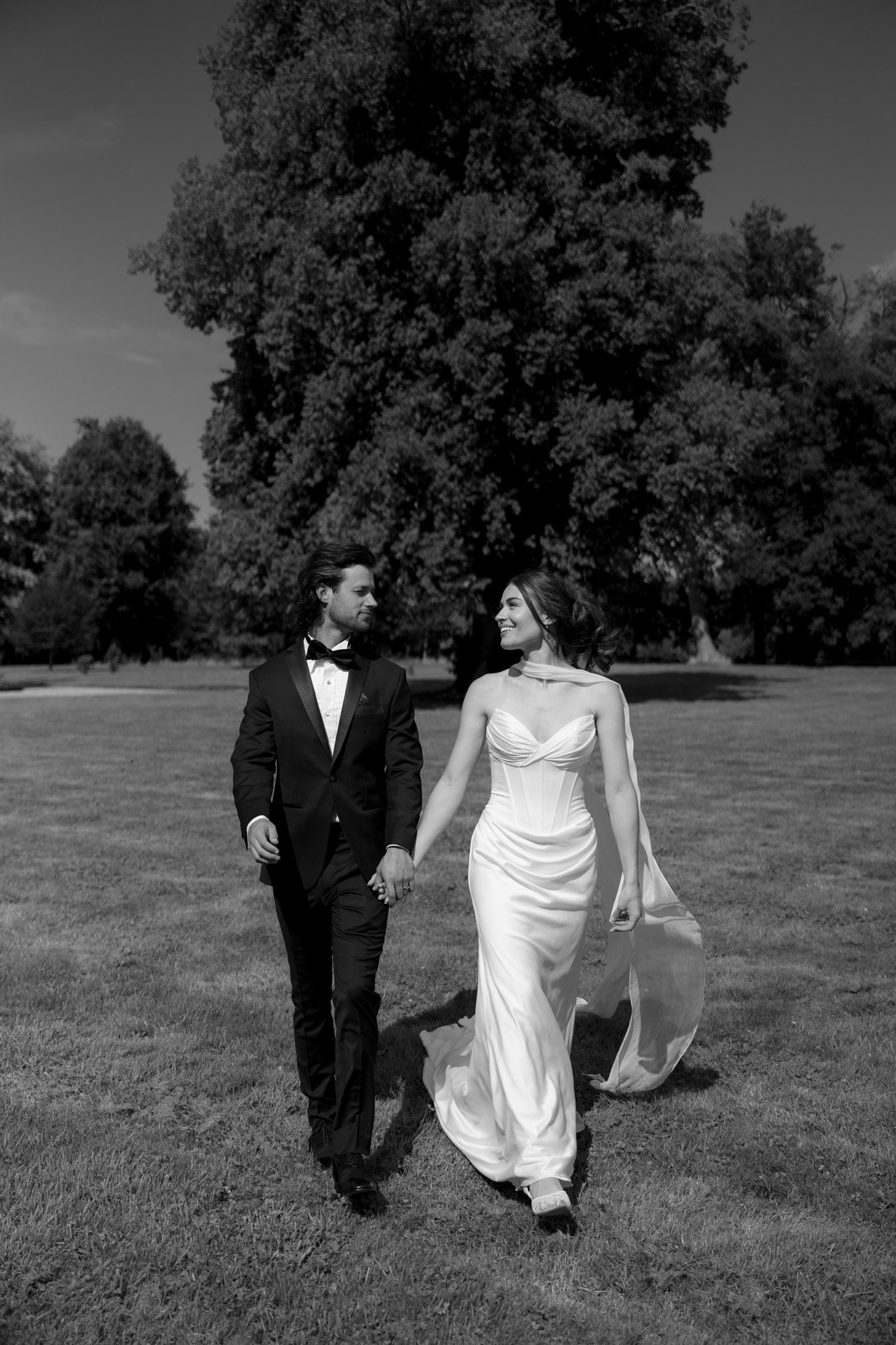 A couple dressed in formal wedding attire, holding hands and walking on grass in a park, with large trees in the background. Normandy Chateau Wedding.