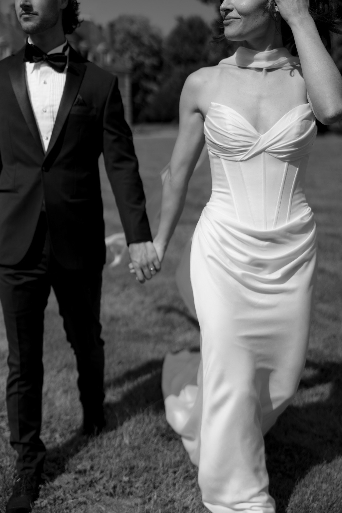 A couple dressed in formal wedding attire hold hands and walk together outdoors on grass.