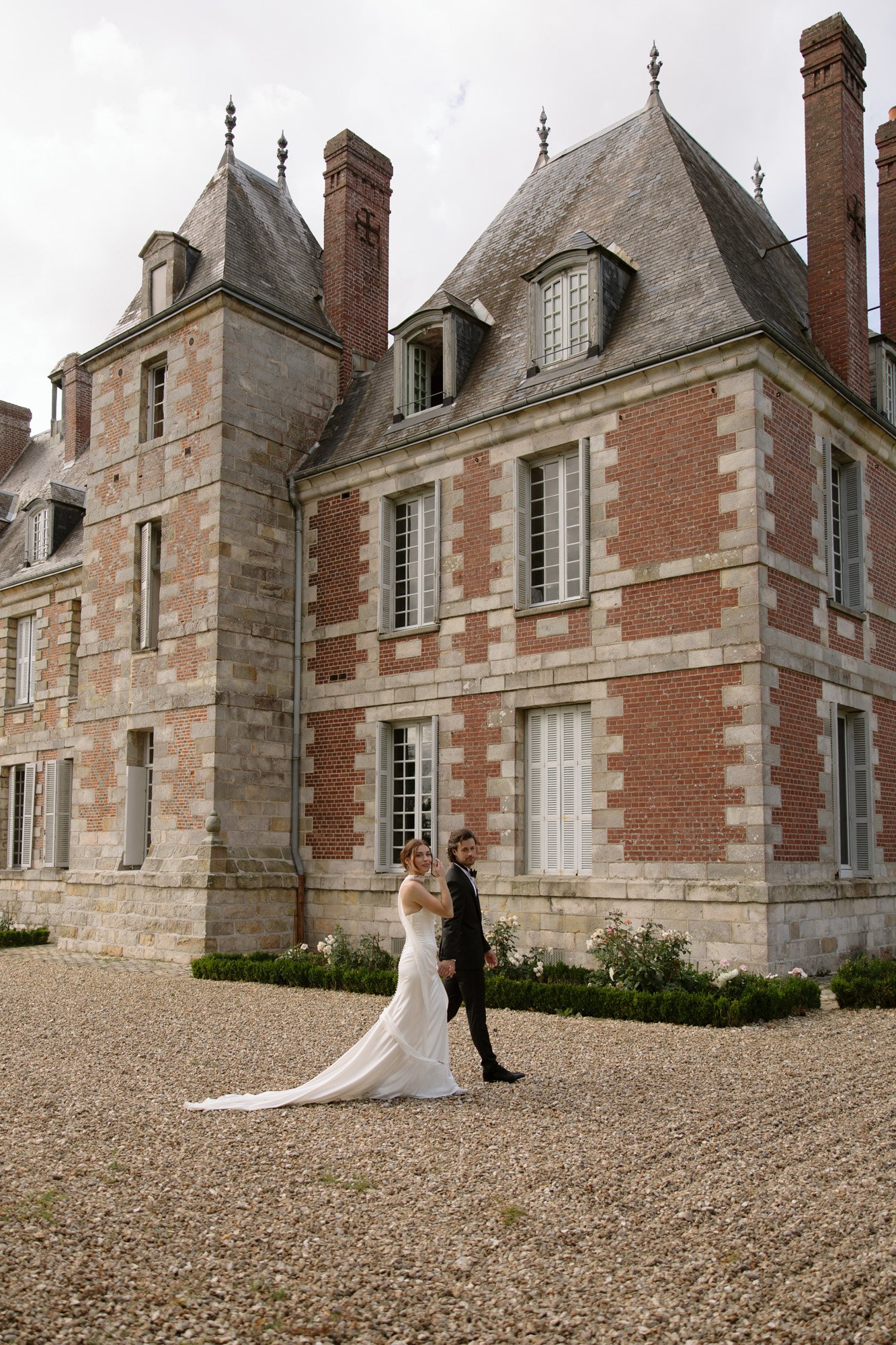 A bride and groom stand together outside a large, historic brick and stone building with tall windows and steep roofs.
