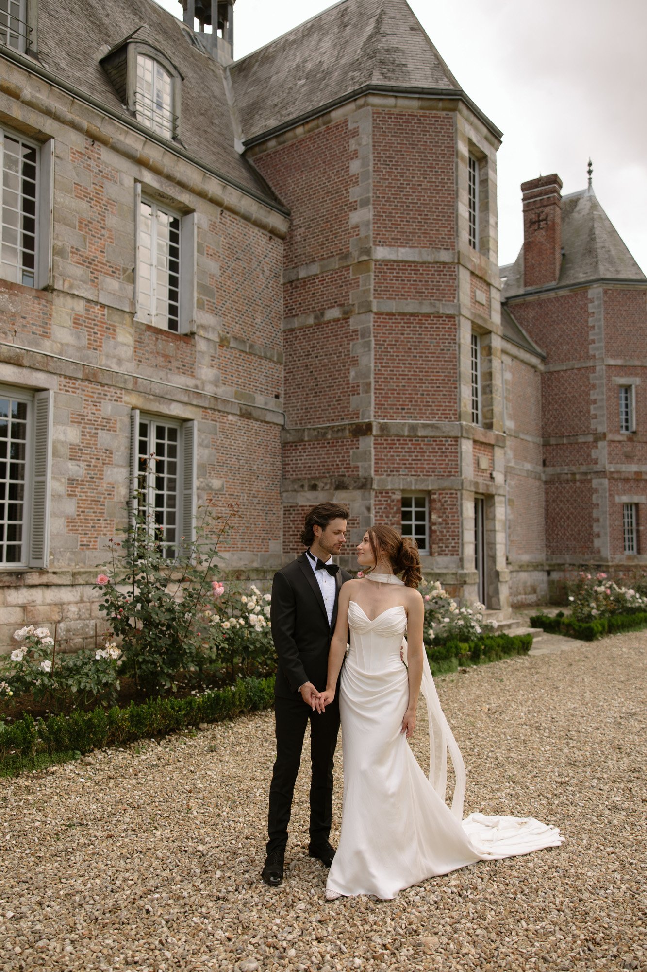 A bride in a white gown and a groom in a black tuxedo stand holding hands outside a large stone and brick building with tall windows.