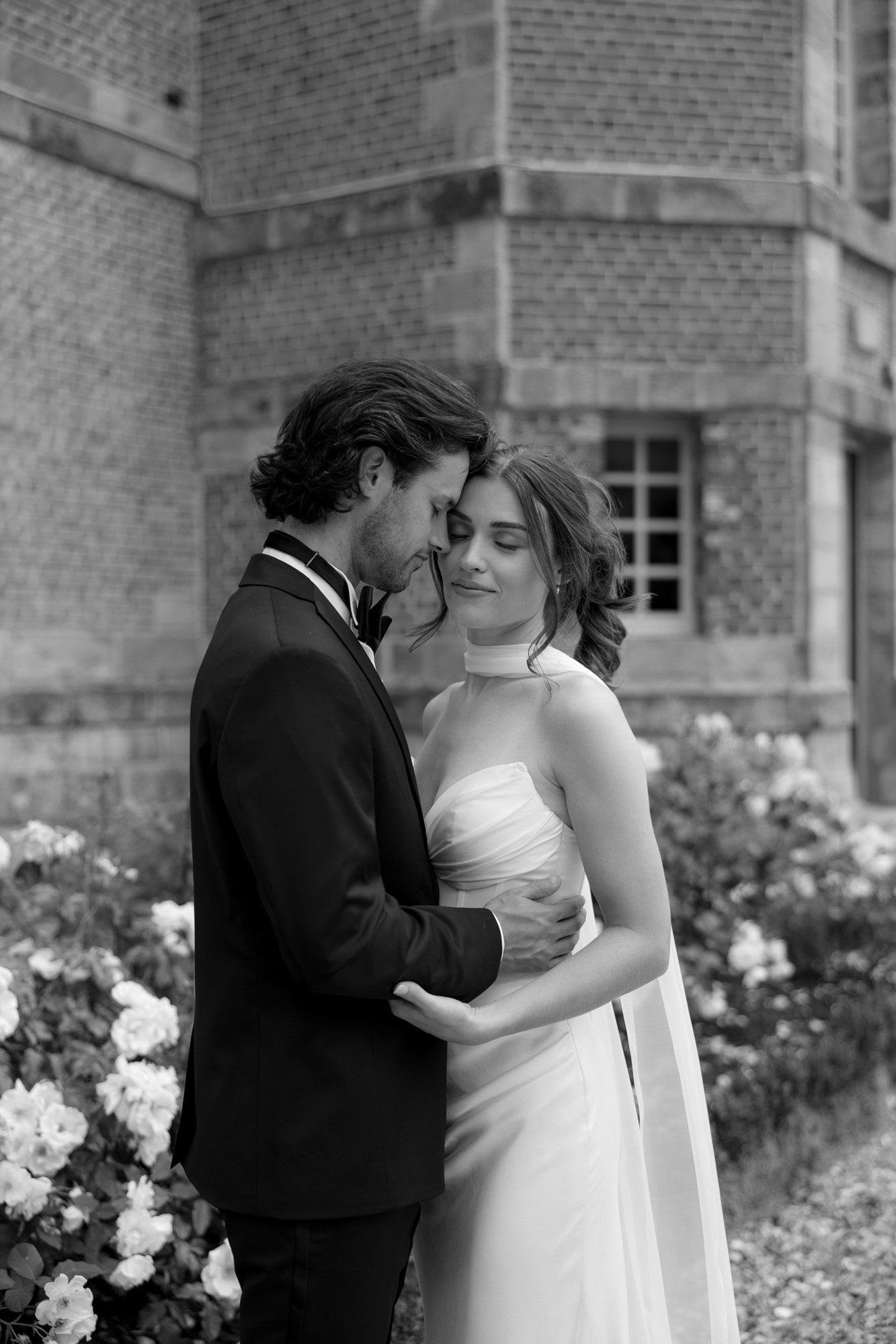 A bride and groom stand closely together outside a brick building, surrounded by blooming flowers. The groom wears a suit, and the bride wears a white gown with a train. Normandy Chateau Wedding.