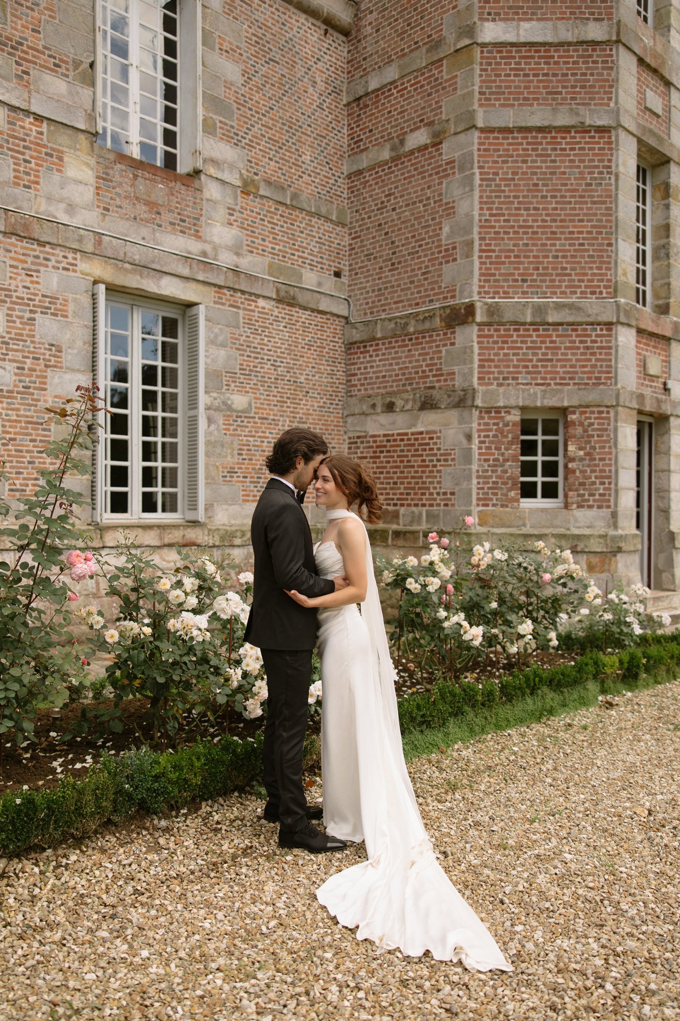 A bride and groom stand close together in front of a brick building, surrounded by blooming roses, on a gravel path.