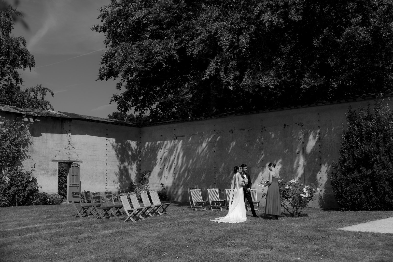 A bride and groom stand with an officiant in an outdoor courtyard with empty chairs, surrounded by trees and a high wall.