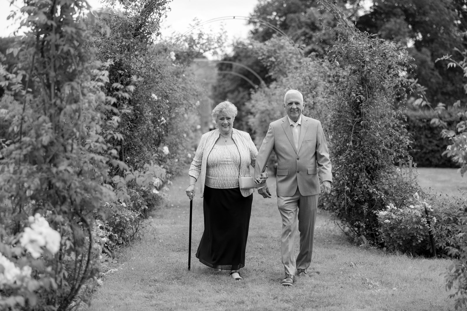An older woman with a cane and an older man walk together through a garden pathway lined with greenery and flowers.