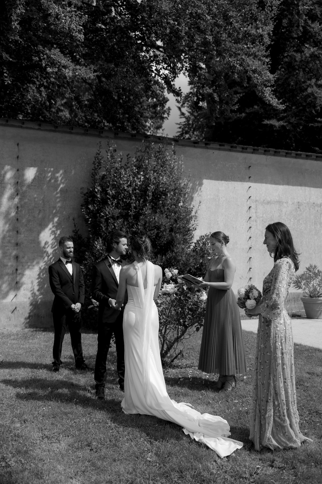 A bride and groom stand facing each other outdoors during a wedding ceremony, accompanied by two women and one man, with trees and a wall in the background. Normandy Chateau Wedding.