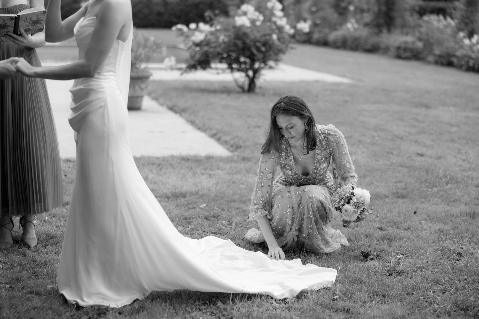 A woman in a gown kneels on the grass, arranging the train of another womans wedding dress in an outdoor garden setting.