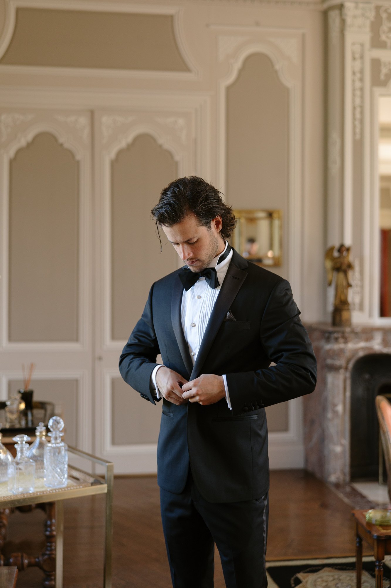 A man in a black tuxedo buttons his jacket in an elegantly decorated room with ornate walls and a marble fireplace. Normandy Chateau Wedding.