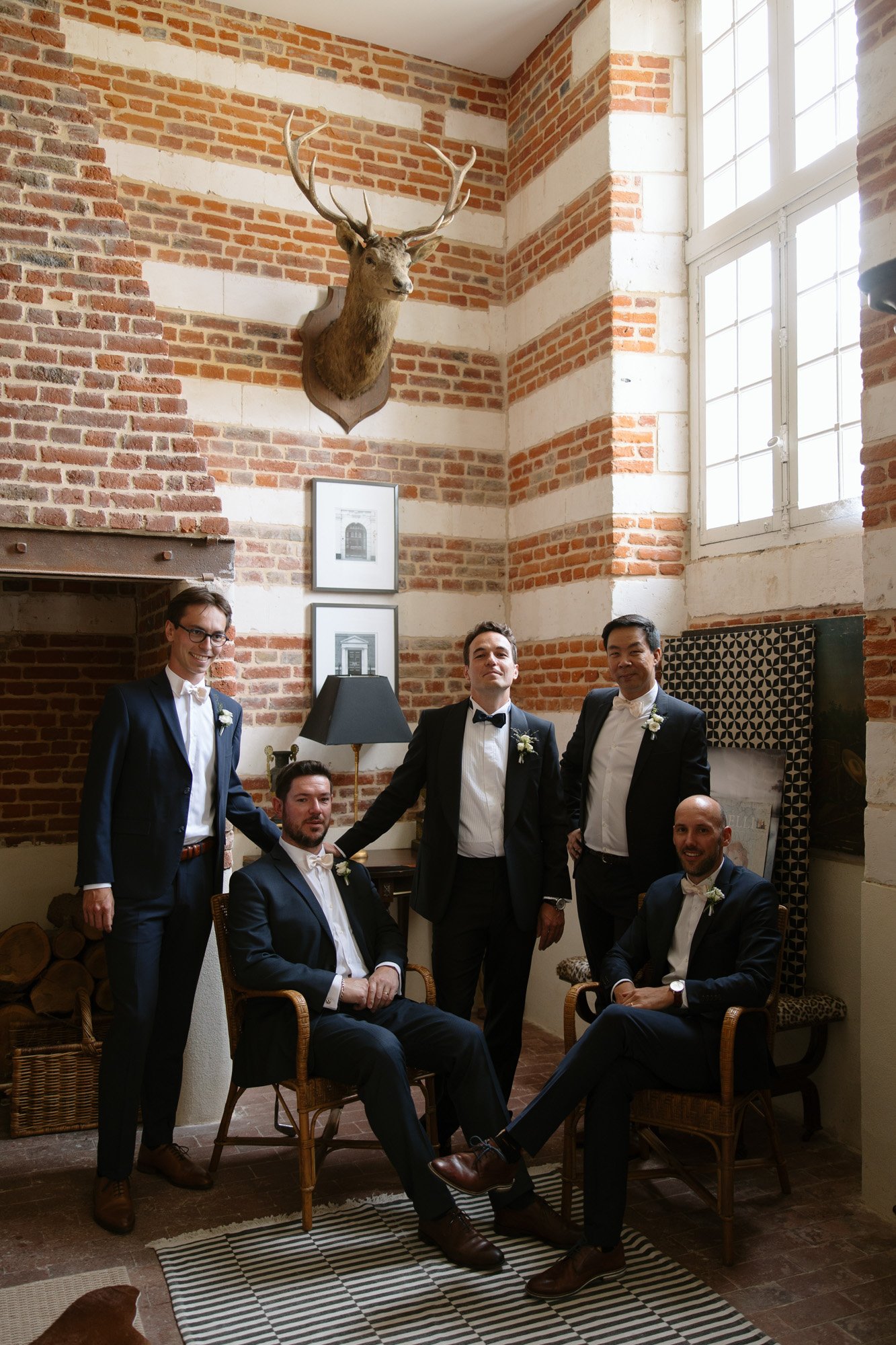 Five men in suits pose for a group photo in a brick room with a fireplace, mounted deer head, and large window; two are seated, three are standing.