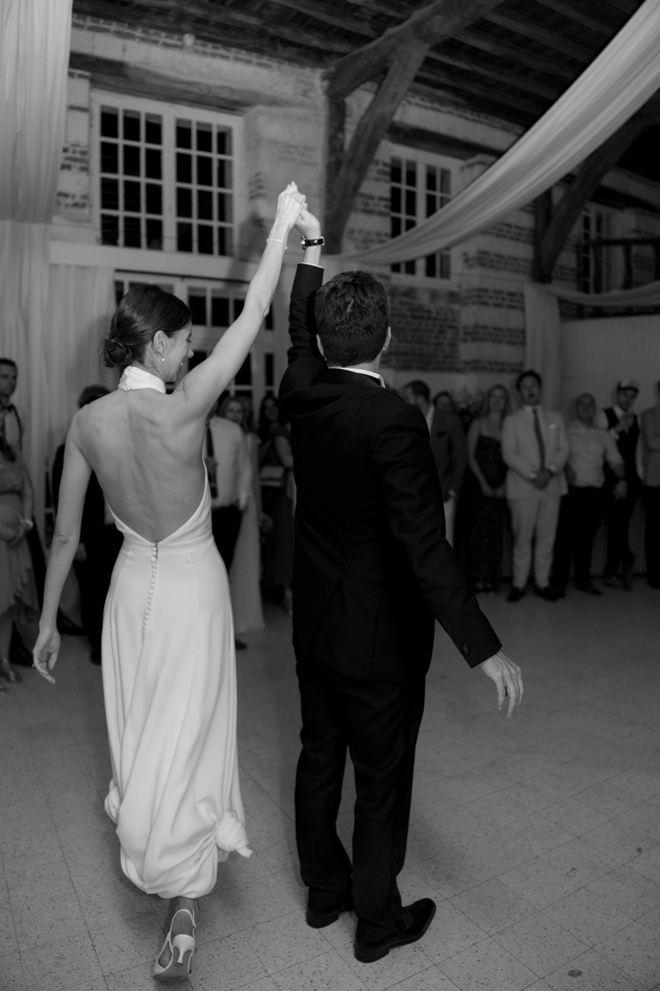 A bride and groom, seen from behind, hold hands and raise their arms while standing in front of a group of guests at an indoor wedding reception. Captured by Northern France wedding photographer.