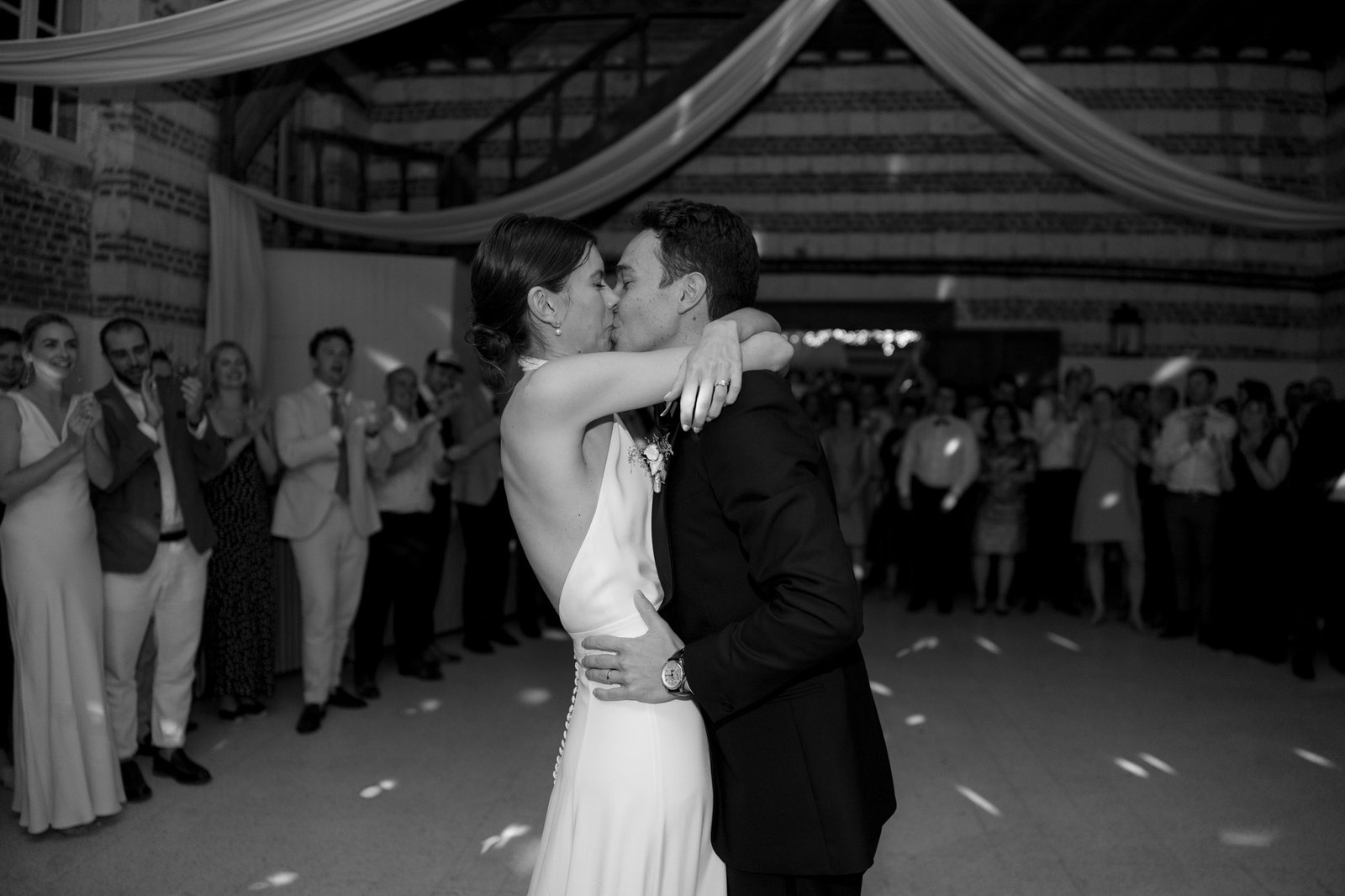 A couple shares a kiss while dancing in the center of a room, surrounded by a crowd of people clapping and watching.
