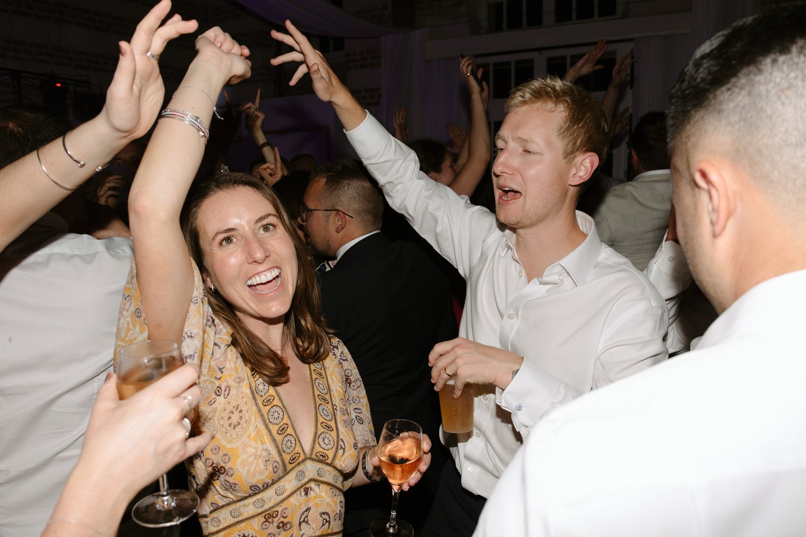 A group of people dressed in semi-formal attire are dancing and holding drinks at an indoor party or celebration.