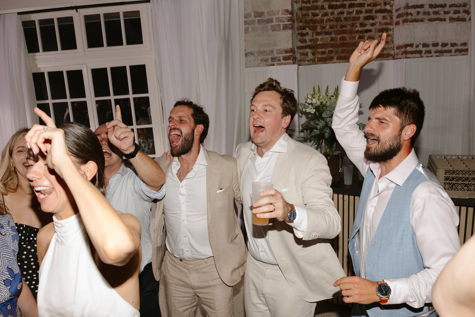 A group of people dressed in formal attire are cheerfully singing and raising their arms at an indoor event or party.