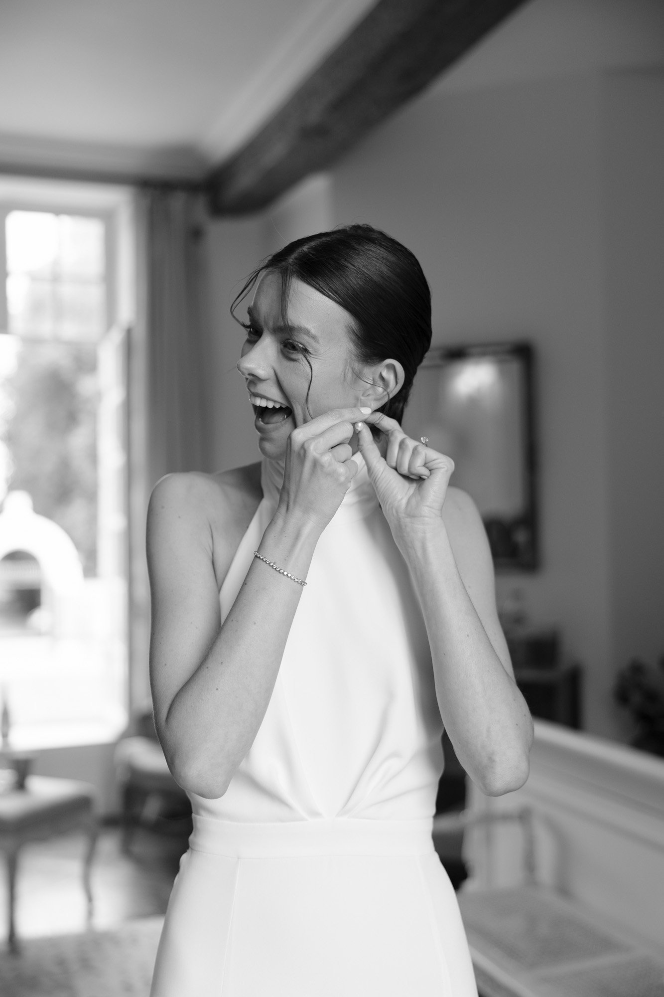 A woman in a sleeveless dress smiles while putting on an earring in a well-lit room with a window in the background.