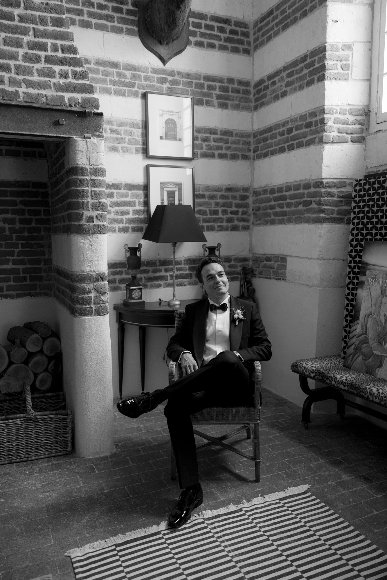 A man in a tuxedo sits on a chair in a brick-walled room with framed art, a lamp, and logs beside a fireplace.