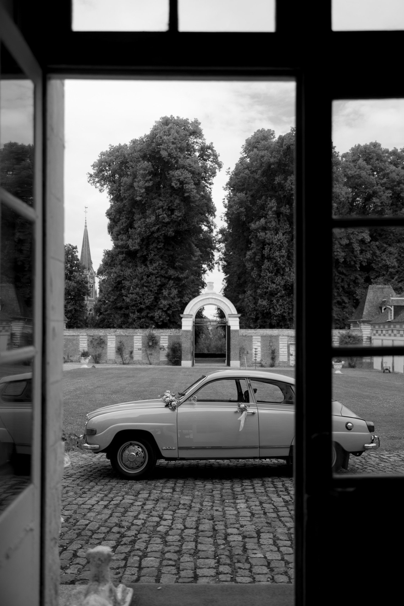 Black-and-white photo of a vintage car with floral decorations parked on cobblestones, viewed through a doorway, with trees and a stone archway in the background. Captured by Northern France wedding photographer.