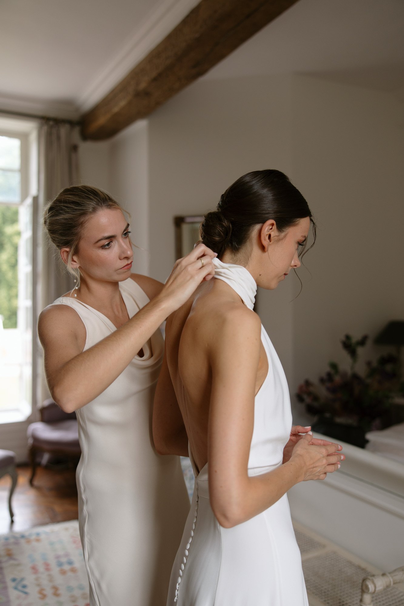 A woman helps another woman fasten the back of her white dress in a softly lit room with wooden beams and a window in the background.