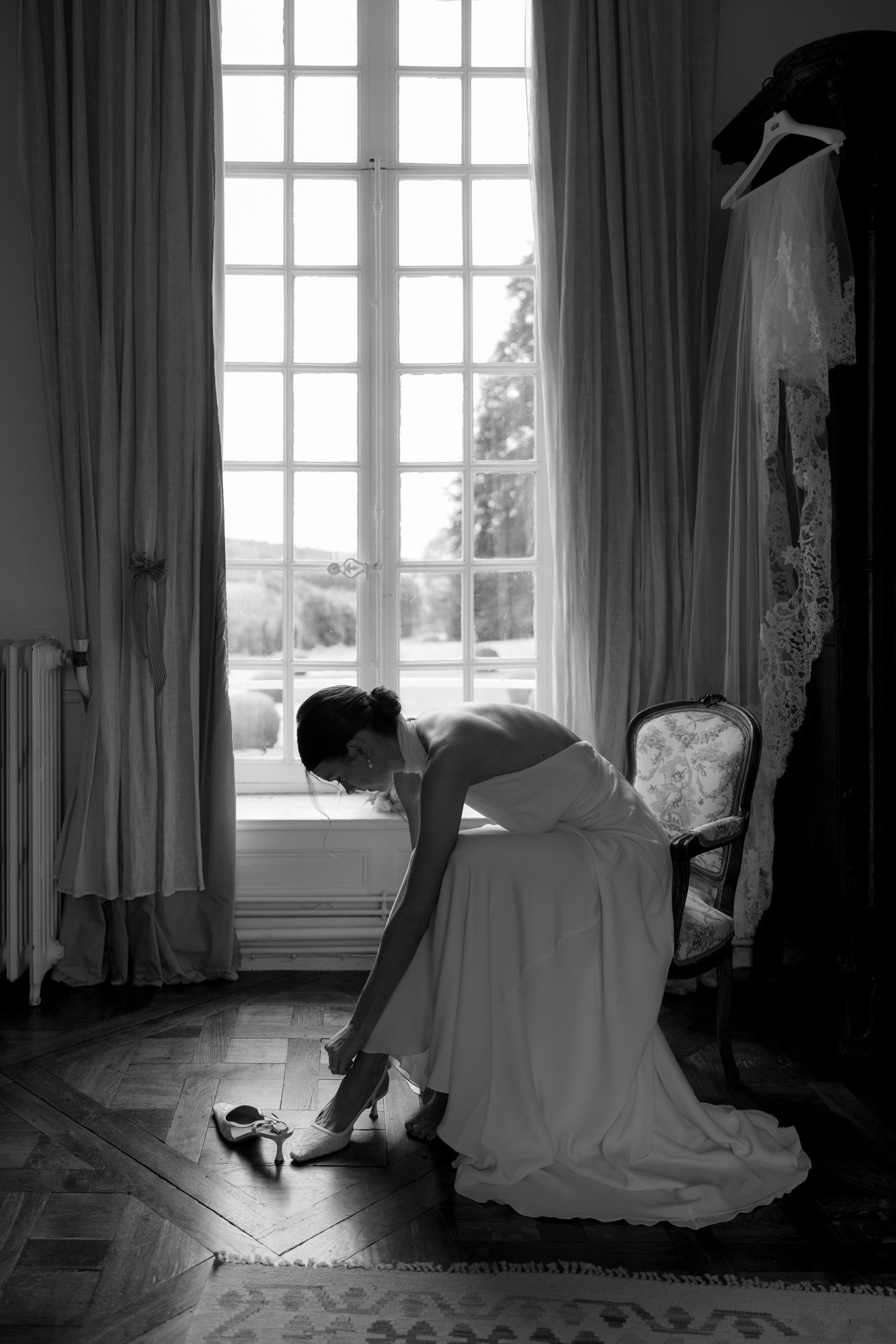 A bride in a white dress sits on a chair by a window, bending down to put on her shoe; a wedding veil and dress hang nearby.