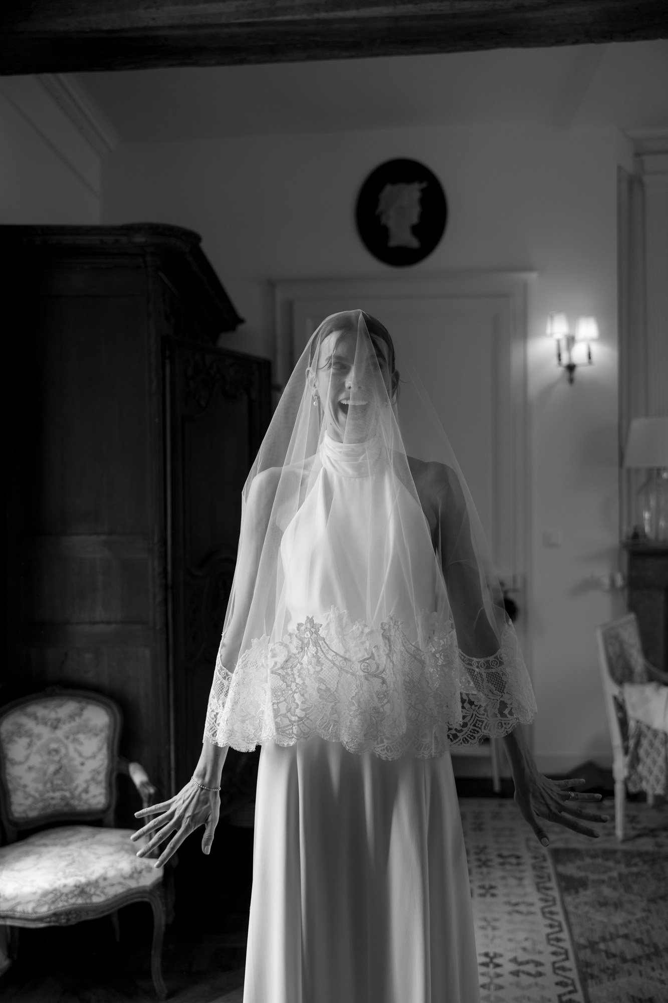 A bride in a lace-trimmed veil and gown stands indoors with her mouth open and arms outstretched, surrounded by vintage furniture.